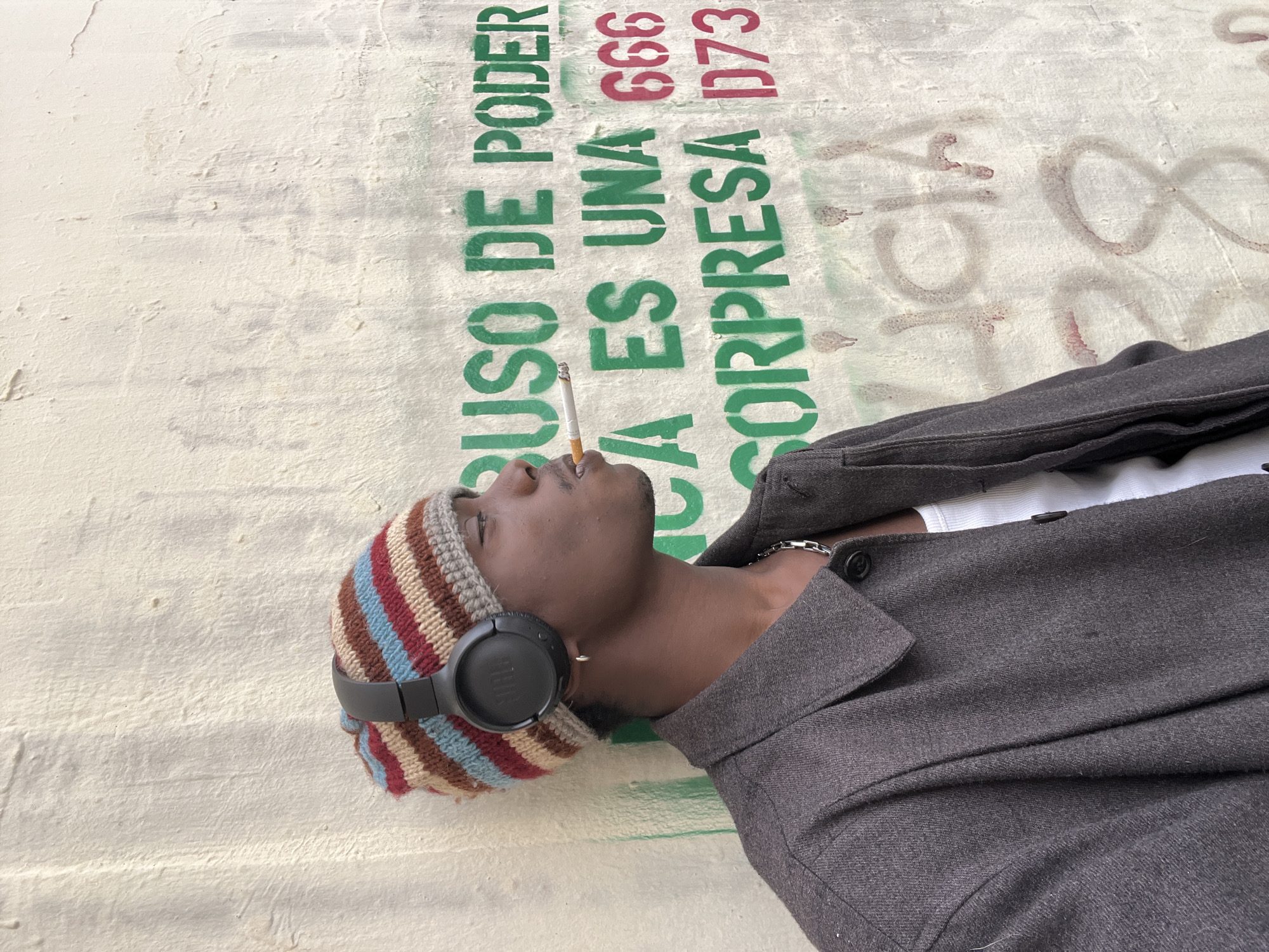 A person is depicted in a close-up shot, lying down or leaning against a textured, light-colored wall. The individual appears to be male with dark skin, wearing a multicolored striped knit beanie (featuring shades of brown, blue, red, and cream) and black over-ear JBL headphones. He is dressed in a dark grey or charcoal-colored jacket over a white undershirt, with a silver-colored chain visible around his neck.
The person is actively smoking a cigarette, which is held between his lips, and he is looking upwards. His expression suggests a moment of relaxation or contemplation.
The background is dominated by a cream or off-white wall with visible texture and signs of wear. Prominently displayed on the wall is green stenciled graffiti in Spanish, which reads: "ABUSO DE PODER" (Abuse of Power) and "ES UNA SORPRESA" (is a surprise). Above parts of the green text, there are red stenciled numbers: "666" and "D73". Faint, older reddish-brown graffiti is also visible in other areas of the wall, partially obscured.
The bright lighting indicates that the scene takes place during the daytime. The Spanish graffiti is consistent with the provided location context of Barcelona, Spain, suggesting an urban setting.
