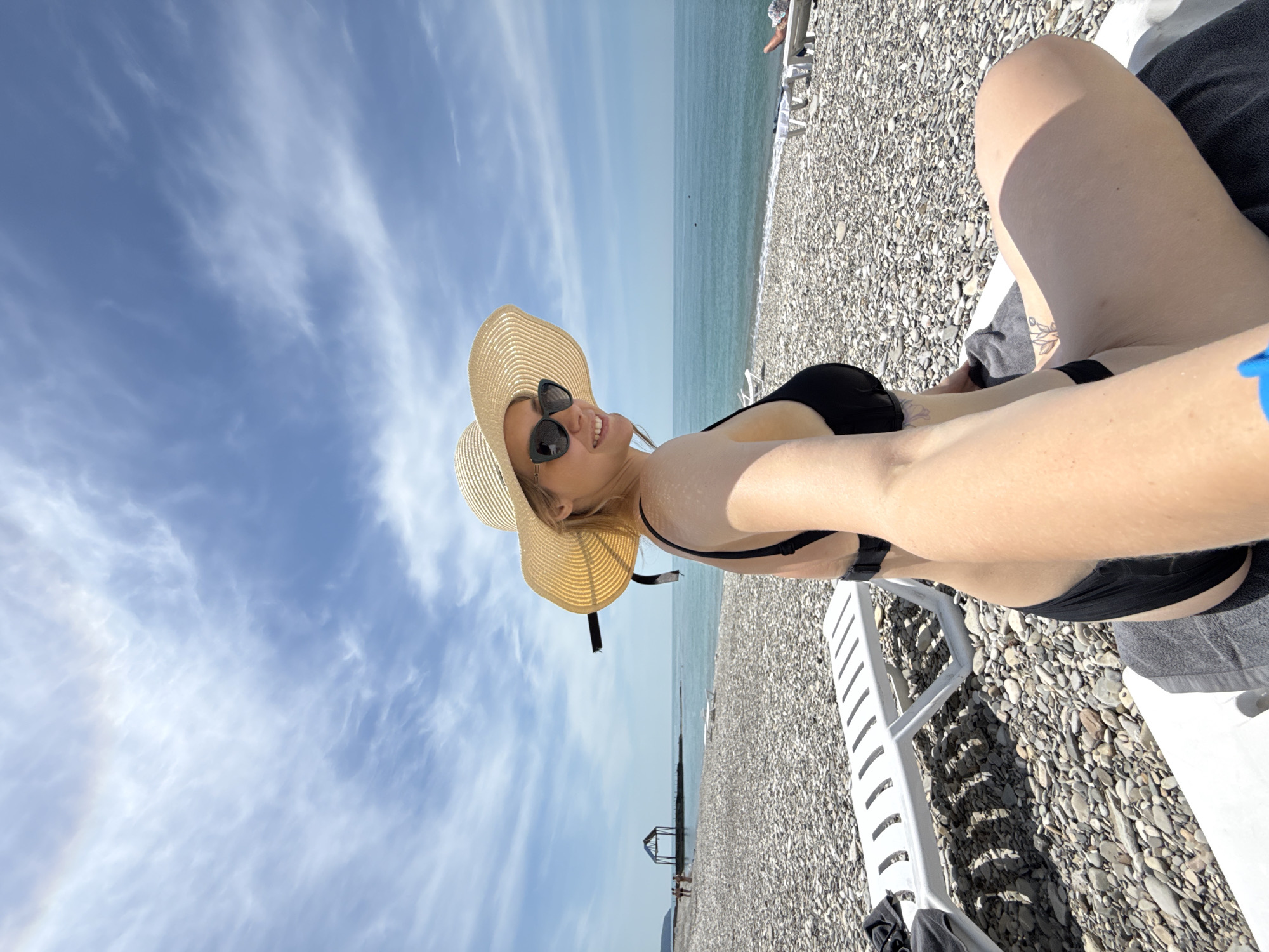 A young woman is depicted taking a selfie on a pebble beach in Nebug, Russia, on a sunny day. She is positioned in the foreground, facing towards the right side of the frame while looking back at the camera with a smile. She wears a wide-brimmed straw hat and dark sunglasses, a black bikini top, and black bikini bottoms. Her right arm is extended, holding the camera, and her left shoulder and arm show visible wetness or glistening from water. Tattoos are visible on her inner left arm and upper left thigh. She is seated on a white plastic lounge chair covered with a dark grey towel.
The beach foreground consists entirely of light and dark grey pebbles of varying sizes. White plastic lounge chairs are scattered across the beach, with one partially visible to her left and others further down the shore. The calm, light blue-green sea extends across the midground to the horizon, meeting a bright blue sky with scattered white, wispy clouds. In the distance to the left, a dark-roofed, open-sided structure, possibly a pier or pavilion, extends into the sea, with indistinct human figures visible near it and in the water. Faint landforms or mountains are visible along the distant coastline.