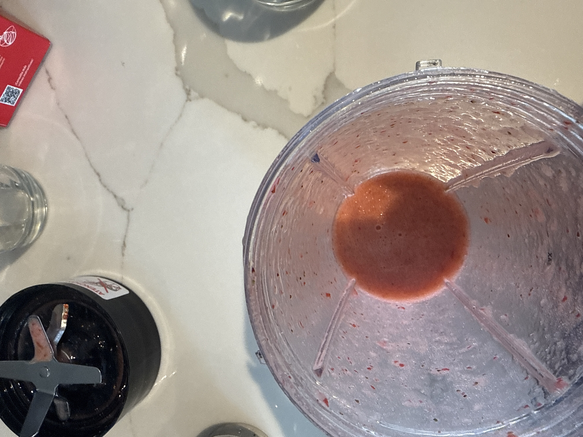 The image presents an overhead view of a kitchen setting. The countertop is a light-colored material with subtle patterns suggesting stone or marble. On the left side, a red object, possibly a package or card, partially enters the frame. Adjacent to it is a small glass container, possibly containing a liquid. Further down, a black blender base with a multi-blade attachment is visible.
The primary subject of the image is a transparent blender container, partially filled with a reddish-orange liquid, likely a smoothie or blended mixture. The inside of the container shows remnants of previous blending, with splatters and residue visible on the walls. The liquid forms a concentrated pool at the bottom of the container.
There are no people visible in the image. The objects suggest a domestic setting, likely a kitchen, where food preparation occurs. The scene portrays a blending process in progress or recently concluded. The image is captured with an overhead perspective.
There is no information in the image that indicates a location in the city of San Bernardino Tlaxcalancingo, Mexico. The scene seems to be an indoor location that could be anywhere with similar tools and settings.