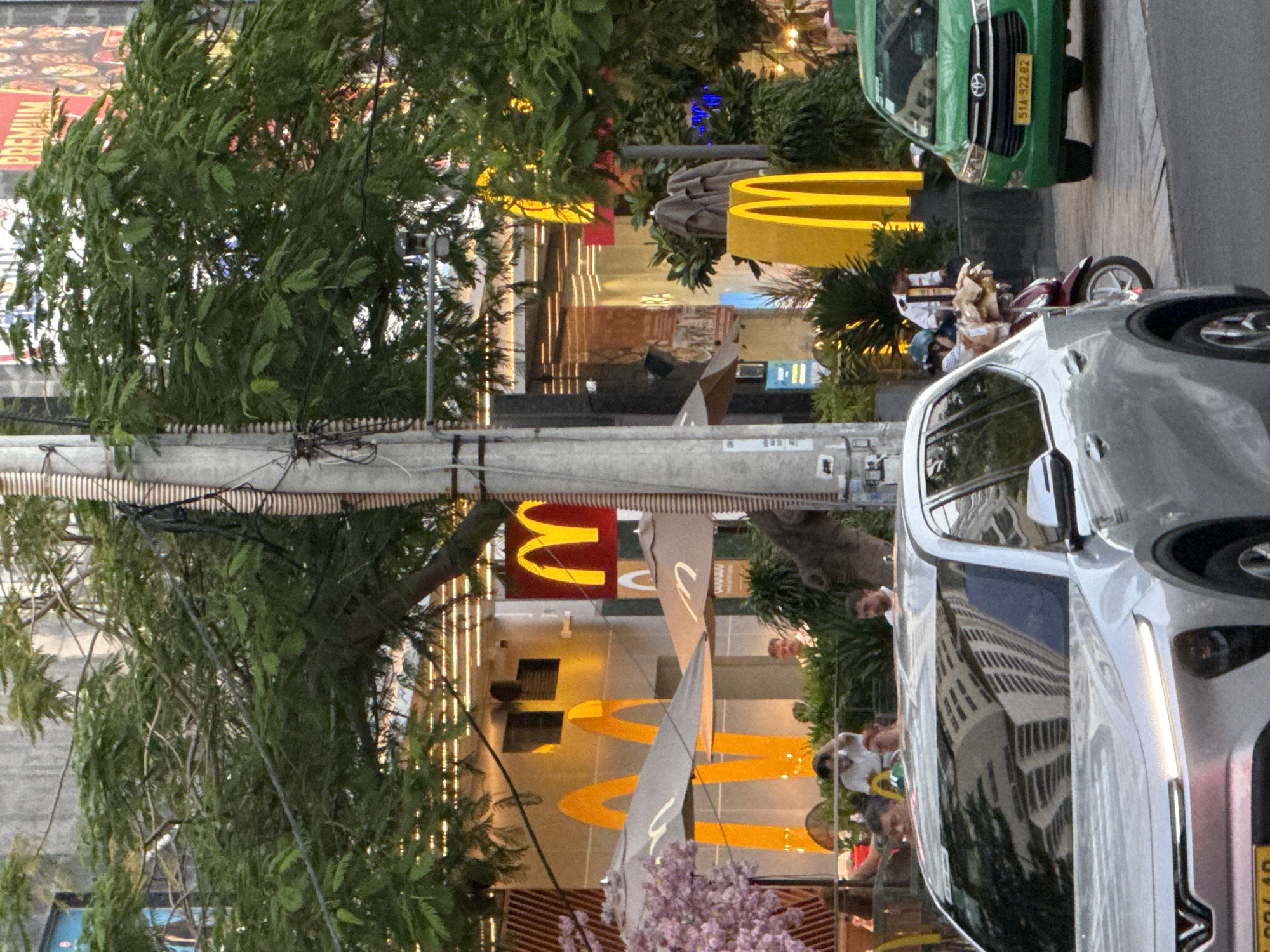 This is a medium shot of a street scene in Nha Trang, Vietnam, likely taken during daylight. The foreground is dominated by the front of a silver car, with its headlights illuminated. To the right, a green taxi with license plate "51A-522.62" is parked on the right side of the road. Behind the taxi, a person on a scooter is stopped, facing away from the camera.
The mid-ground features a McDonald's restaurant with its distinctive golden arches prominently displayed on the building and as large standalone signs. Several tables with umbrellas are set up outside the restaurant, with a few patrons seated. A tree with pink blossoms is visible to the left of the McDonald's entrance.
The background shows the multi-story buildings lining the street, one of which has a large "PREMIUM" sign at the top. Overhead, trees with green foliage obscure parts of the buildings and signage. Utility poles and wires are visible throughout the scene. The overall impression is of a busy urban commercial area.