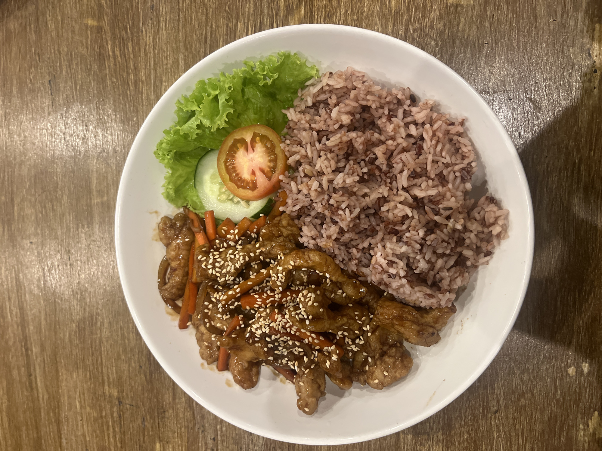 A top-down, close-up shot depicts a white, round plate filled with food on a wooden surface. The plate contains a portion of red rice at the upper right quadrant. Adjacent to the rice, at the upper left quadrant, is a lettuce leaf with a tomato slice and a cucumber slice. At the bottom left quadrant, there is meat covered in glaze sauce with sesame seeds, mixed with carrots. The scene is located in Tibubeneng, Indonesia. There are no people or interactions visible.