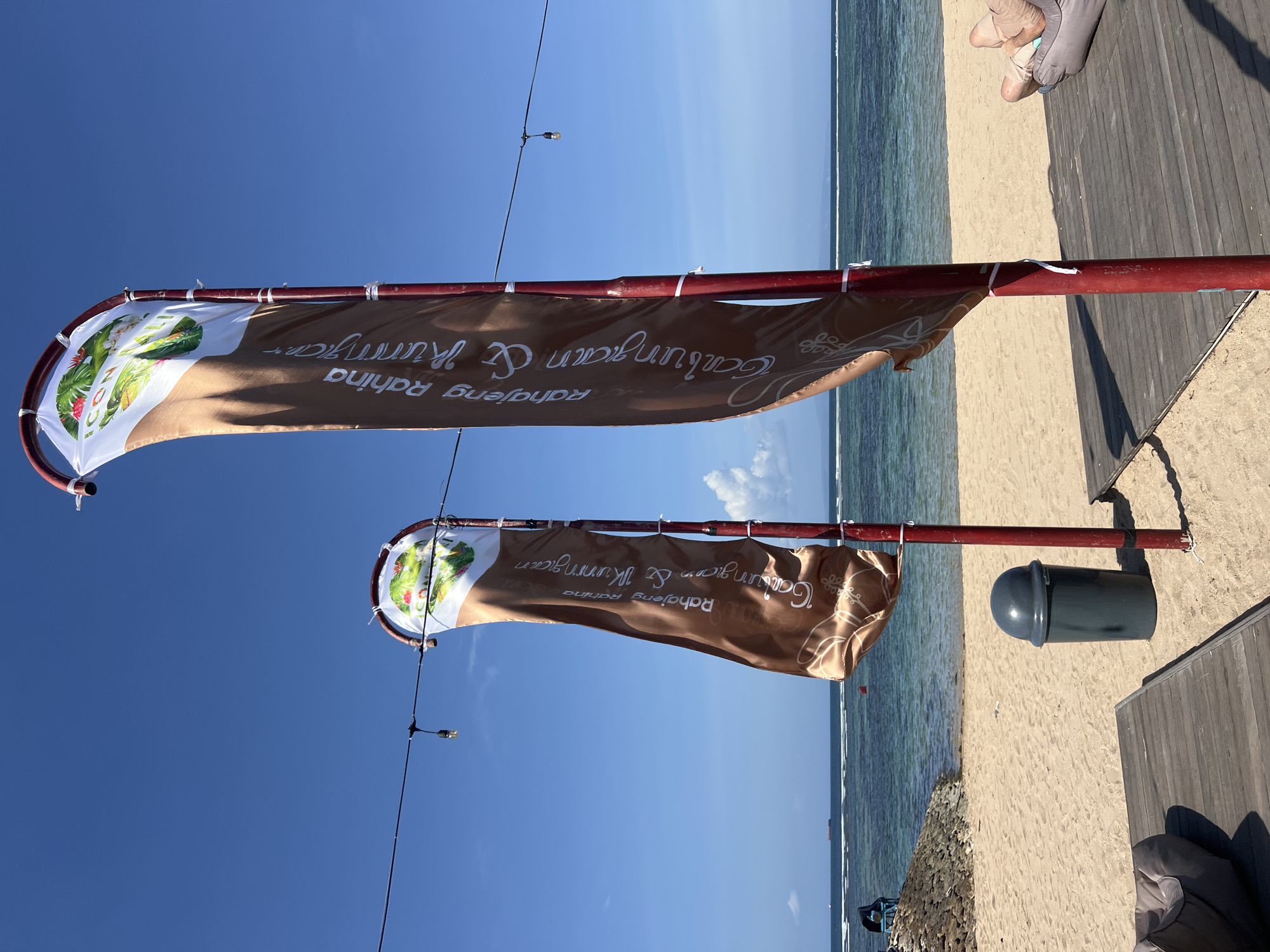 The image shows two banners displaying the text "Gaiulian Rauhahag & Kummgar". The banners are attached to red poles and are set against a clear blue sky. The top banner features a circular design with images of tropical plants and what appears to be a salad. The bottom banner has a similar design but is partially obscured.
In the background, a sandy beach stretches towards the ocean, with calm, blue water. A wooden boardwalk is visible on the right side of the image, with a gray trash bin nearby. In the distance, a person is partially visible, reclining on the boardwalk. The scene suggests a bright, sunny day, possibly in the morning or afternoon, given the clear sky and the lighting on the beach and water. The location appears to be a tropical beach resort or a similar establishment. The overall atmosphere is relaxed and serene, typical of a vacation setting.