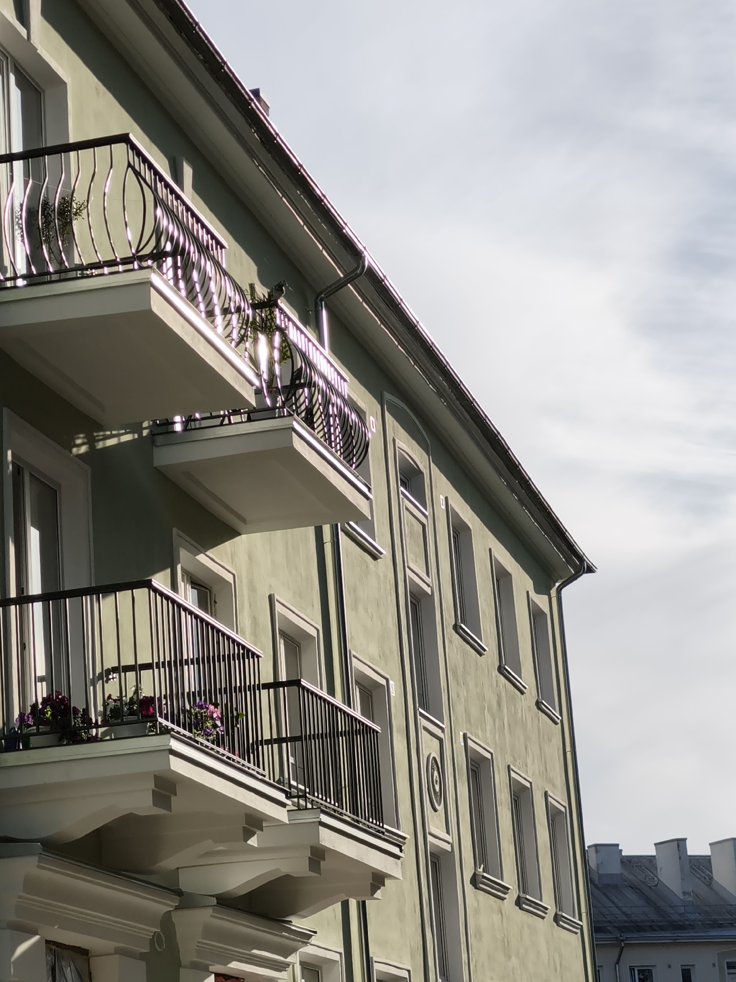 An upward angled exterior shot of a light green apartment building with multiple balconies, located in the city of Tallinn, Estonia. The building has a textured facade and cream-colored window frames. Two prominent balconies are visible in the foreground, each featuring black metal railings and decorative architectural elements underneath. Potted plants with purple and pink flowers are placed on the railings of the lower balcony. Sunlight reflects off the metallic surfaces and the building's facade. The sky above is overcast with a bright, hazy appearance. In the background, the roofs of other buildings can be seen. No people are present in the image.