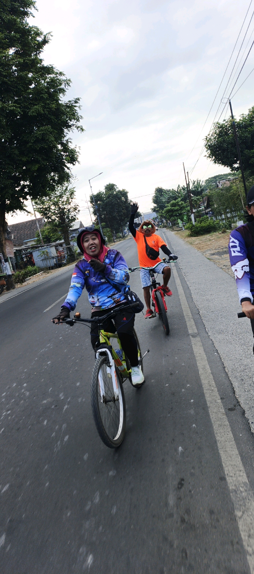 The image depicts a group of three individuals cycling on a paved road in Kasiyan, Indonesia. The foremost cyclist, on the left, is a woman wearing a pink headscarf, a blue and purple long-sleeved shirt with a colorful pattern, black pants, and white sneakers. She has on black gloves and a helmet, and is looking towards the camera with a wide smile and her mouth open. She is riding a green and white bicycle. To her right and slightly behind is a man wearing an orange long-sleeved shirt, light blue shorts, and a fanny pack across his chest. He has sunglasses on and is waving enthusiastically with his right hand, also wearing a helmet. He is riding a red bicycle. On the far right, partially visible, is a third person wearing a dark long-sleeved shirt with white accents, riding a bicycle. The road appears to be a two-lane street with a white dividing line. The environment includes trees and buildings lining the side of the road, consistent with a semi-urban or suburban setting. The sky is overcast. The photograph is taken from a low angle, giving a dynamic perspective of the cyclists in motion.