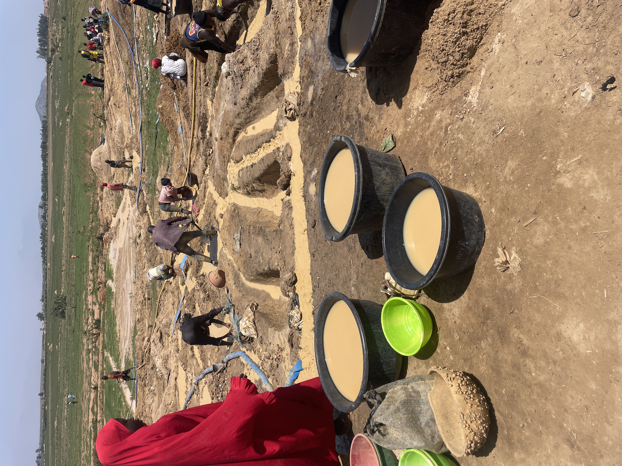 The image shows an outdoor scene in Maiyanga, Nigeria. In the foreground, there are four large black buckets, three filled with a light brown liquid, likely muddy water. There is also a collection of smaller colored bowls and containers nearby, including a bright green bowl. The ground is predominantly bare dirt and sand.
In the background, there are several people engaged in digging or working in the soil. The ground shows a series of narrow trenches or excavations, some filled with muddy water. Several blue pipes and yellow hoses are visible, suggesting water is being used in the digging process. A mix of green vegetation and bare soil stretches to the horizon, where the sky is a light haze.
The people in the background appear to be manually digging and moving material. Some are squatting, while others are standing, possibly transferring soil or water. The presence of buckets in the foreground and the trench-like excavations imply the removal of material from the ground.