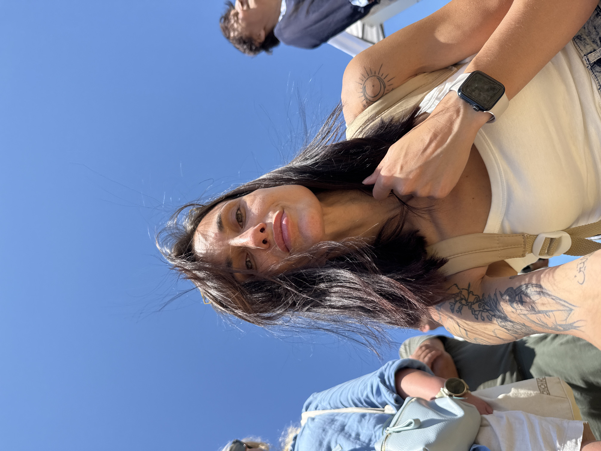 A low-angle outdoor photograph captures a group of people against a clear, bright blue sky in Gigen, Bulgaria. The primary subject is a woman in the foreground, positioned centrally, looking directly at the camera. She has long, dark, windblown hair, light skin, and a slightly pursed-lip expression. She wears a white tank top, with a tan backpack strap visible over her right shoulder. Her left wrist displays a smartwatch with a dark frame and light-colored band. Visible tattoos include a sun and crescent moon design on her right upper arm, and an extensive floral or abstract design on her left shoulder and upper arm, with what appears to be Arabic script on her lower left forearm. Her right hand is raised, with fingers touching her hair near her neck.
To the left of the main subject, a second person is partially visible, wearing a light blue long-sleeved shirt and carrying a light blue shoulder bag. Behind them, a third person in a green shirt is visible. To the right, another person with dark hair, wearing a dark blue shirt, is seen in the mid-ground, out of focus, looking upwards and to the right. The scene is illuminated by bright, natural daylight, indicating an open outdoor environment.