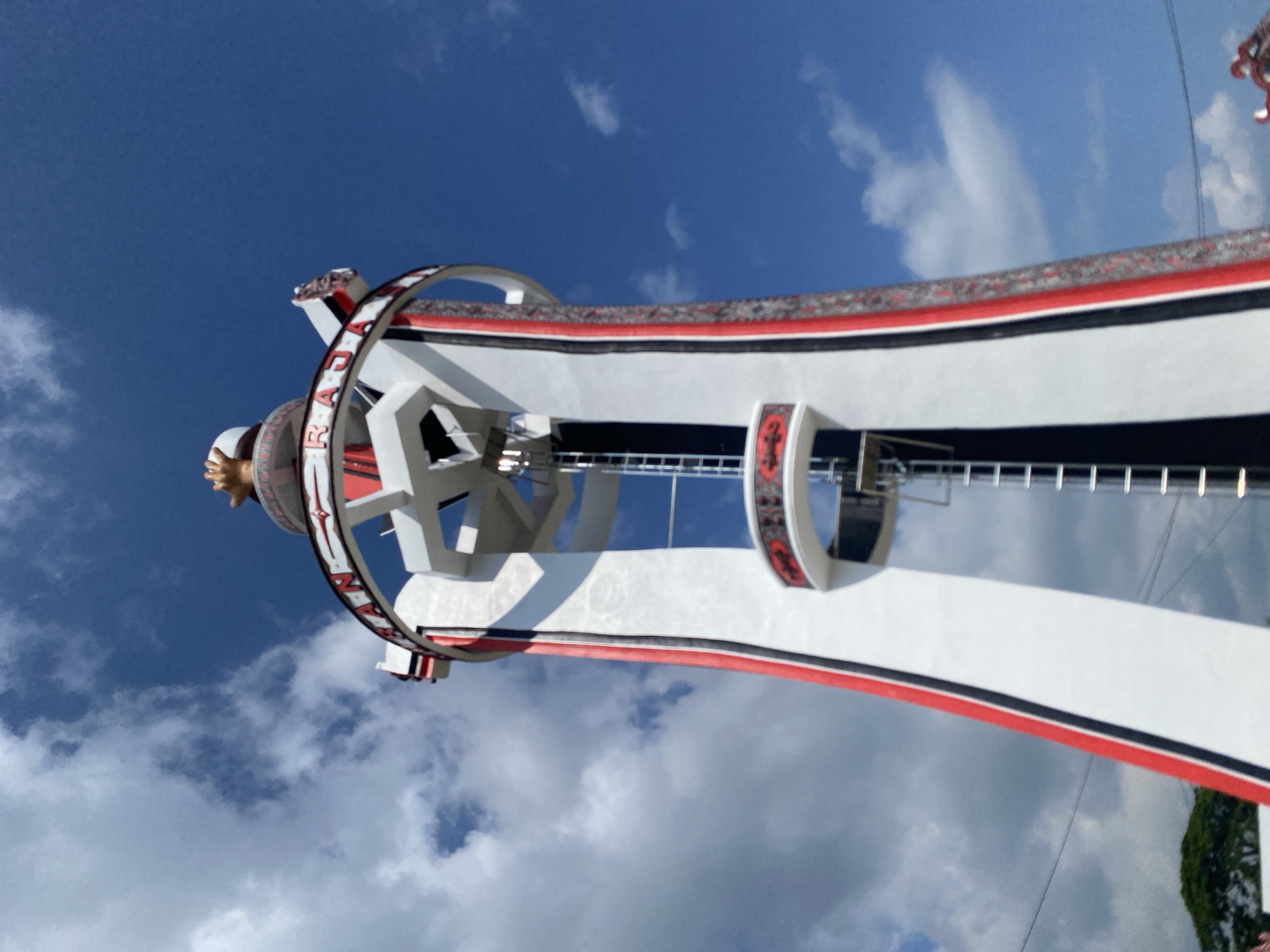 The image shows a low-angle view of a tall, white tower with red and black accents, against a bright blue sky with scattered white clouds. The tower has a conical shape, widening towards the base, and is adorned with vertical red and black stripes along its white surface. A circular band with red and white decorative patterns encircles the upper part of the tower, with what appears to be text written in red on it, reading "RAN RAJA." There is a ladder visible running vertically along the side of the tower, and a small platform with a railing extends from the tower partway up. At the very top of the tower, a golden statue of hands clasped together is visible. The background is a clear blue sky with fluffy white clouds. The scene appears to be outdoors, likely in an open area, given the unobstructed view of the sky and the presence of greenery at the bottom left of the image. This structure is identified as the Monument Simanjuntak, located in Parlondut, a village in North Sumatra, Indonesia.