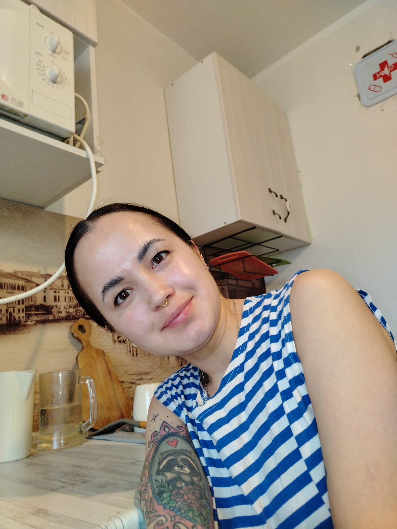 A woman is shown from the chest up, wearing a blue and white striped shirt. She has dark hair pulled back, and a nose ring. She is smiling at the camera. A tattoo is visible on her left upper arm. The background shows a kitchen area with a microwave, a cabinet, a first aid kit, a decorative image of a cityscape (possibly Venice), a wooden cutting board, and various kitchen items. The location is an indoor setting, likely a home kitchen. Noviy, Russia is not identifiable as a specific location within the image.