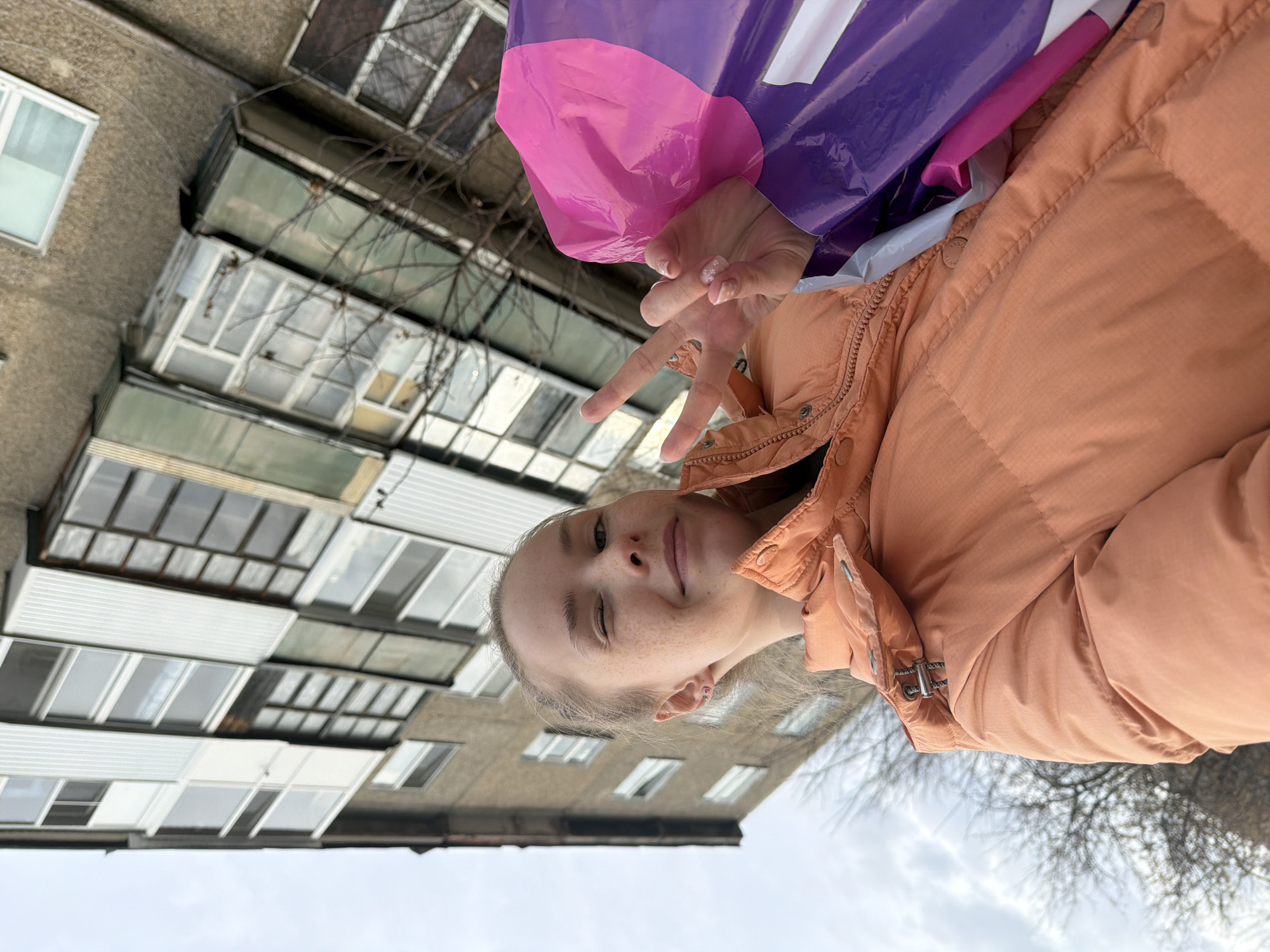 A young person with light hair and freckles is shown in the foreground, winking and making a peace sign with their fingers. They are wearing a light brown puffer jacket and holding a purple and pink plastic bag.
The background consists of a multi-story building with a mix of windows and balconies. There are bare tree branches visible against a gray sky. The photo was taken in Berezniki, Russia.