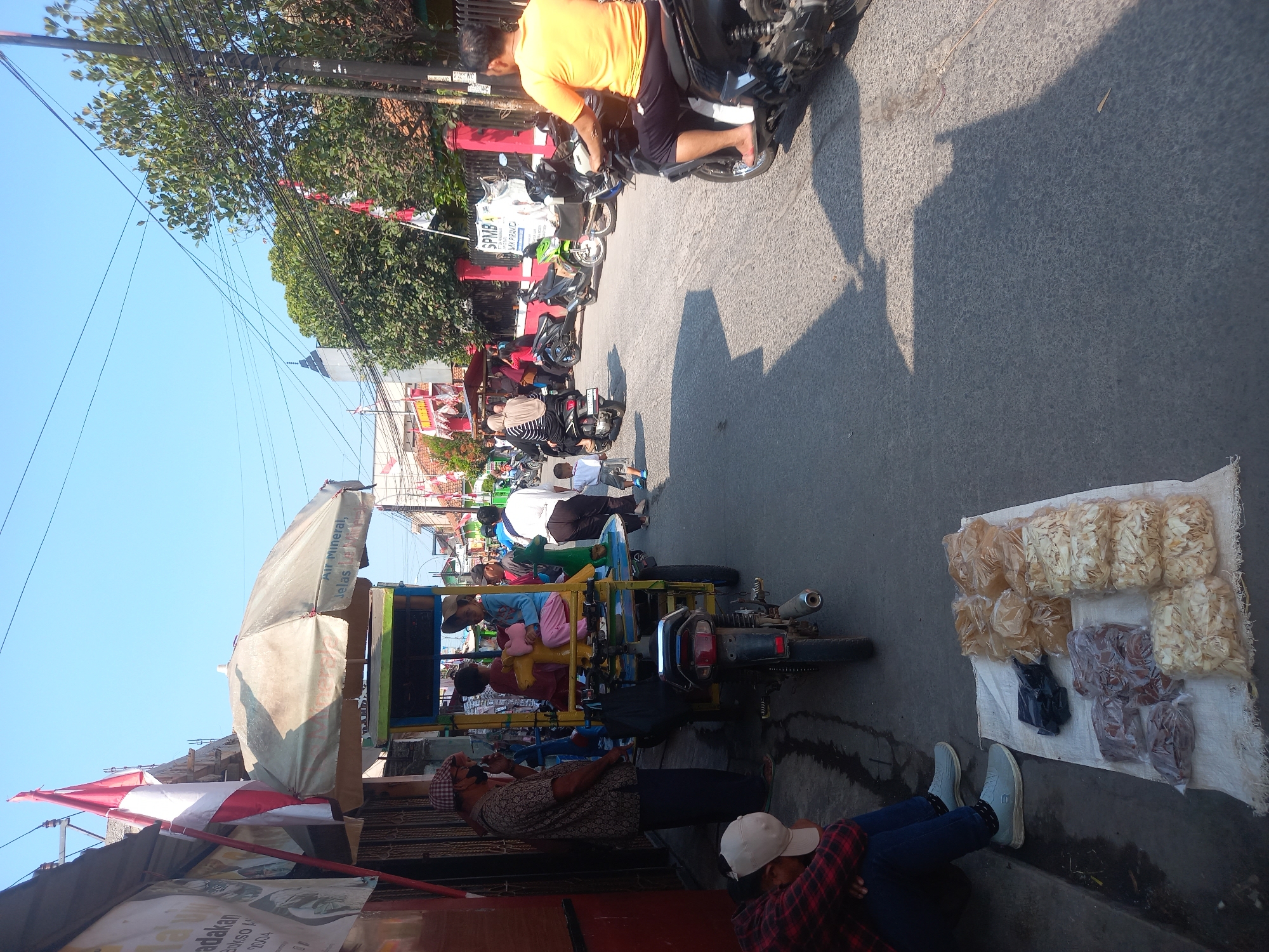 The image depicts a street scene in Linggar, Indonesia, under a clear blue sky. A busy road is present with several motorcycles. In the foreground, on the left side of the road, a person in a red and black plaid shirt and blue jeans is sitting on a curb, looking down. Beside them, a white cloth is spread on the ground with various bagged snacks, possibly for sale. A man wearing a patterned shirt and a checkered cap is standing and conversing with another person, partially obscured by a colorful cart. The cart has an umbrella overhead and appears to be a mobile vending unit, with children inside. Further down the road, a person in an orange shirt rides a motorcycle. Other people and motorcycles are visible in the background, along with buildings and trees. Indonesian flags are also visible, suggesting a local celebration or event.