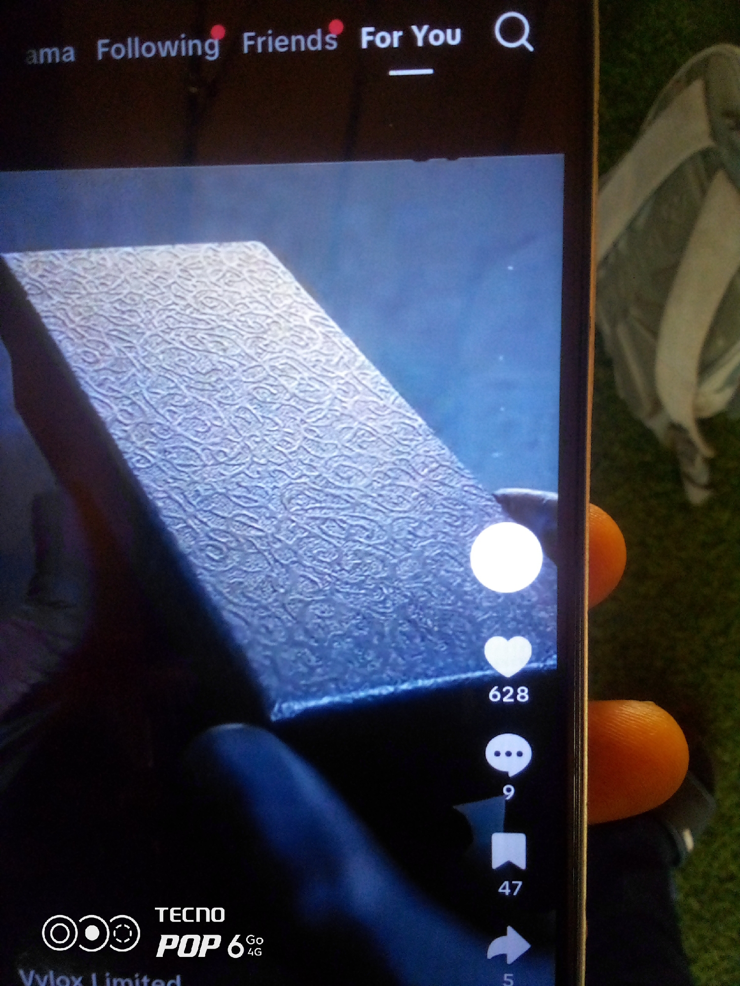 The image shows a close-up of a smartphone screen displaying a video. The video content features a textured box. On the right side of the screen, interactive icons are visible: a heart with the number "628" below it, a speech bubble icon with the number "9" below it, a bookmark icon with the number "47" below it, and an arrow icon with the number "5" below it. In the bottom left corner of the screen, there is a logo with the text "TECNO POP 6 Go 4G" and "Vylox Limited". The background of the image is out of focus and appears to be green grass. A portion of a hand with an orange thumb is visible on the right side of the phone. The top of the phone screen shows navigation elements such as "ama", "Following", "Friends", "For You", and a search icon. The image is taken from a slightly elevated angle. The location in Howayi, Nigeria is not discernible from the provided image.