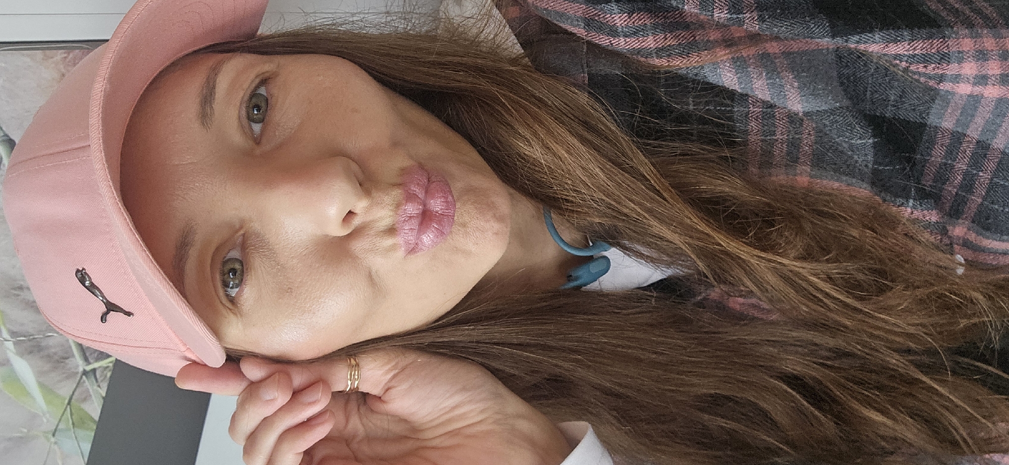 The image shows a close-up, selfie-style portrait of a woman wearing a pink baseball cap with a Puma logo. She has long, brown hair and is making a puckered-lip expression. She is also wearing a gold ring on her right hand, which is near her face. A dark and pink plaid shirt is visible on her. Blue earbuds are around her neck. The background is blurred and appears to be an indoor setting with some plant-like elements. There is no discernible location within the city of Vitsebsk, Belarus.