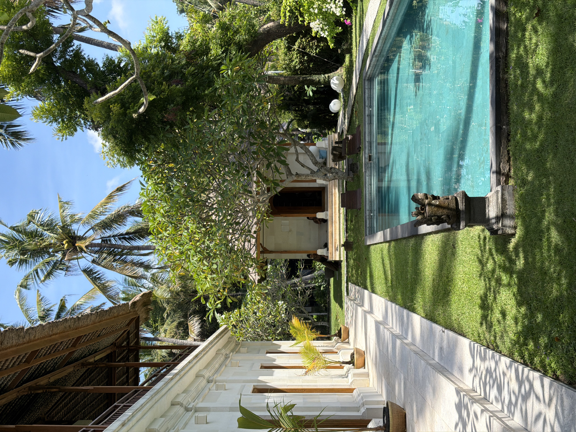 The image shows an outdoor scene, possibly a backyard in Karangasem, Indonesia. The view includes a swimming pool with turquoise water, surrounded by green grass. A stone pathway runs alongside the pool. Trees and various plants provide a lush, tropical environment. The architecture features a white building with a thatched roof and a wooden structure that resembles a porch or gazebo. No people are clearly visible. The overall impression is one of a tranquil, well-maintained outdoor space.