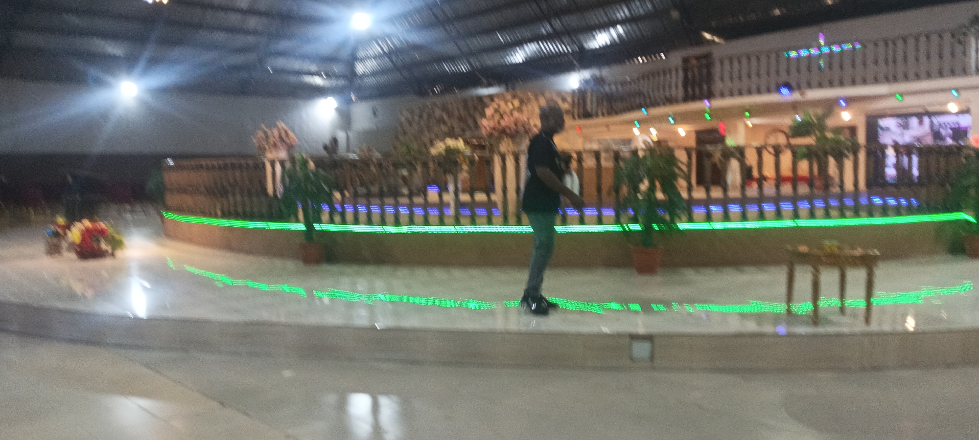The image shows the interior of a large room in a building in what appears to be Rukuba Road, Nigeria. The room is decorated with green and blue lighting accents. A man is walking across the raised platform in the room. There are plants in pots and flower arrangements placed around the perimeter. A small wooden table is also visible. The room appears to be a hall or event space, possibly for a wedding or other celebration. A mezzanine or balcony level is visible in the background, also decorated with lights.