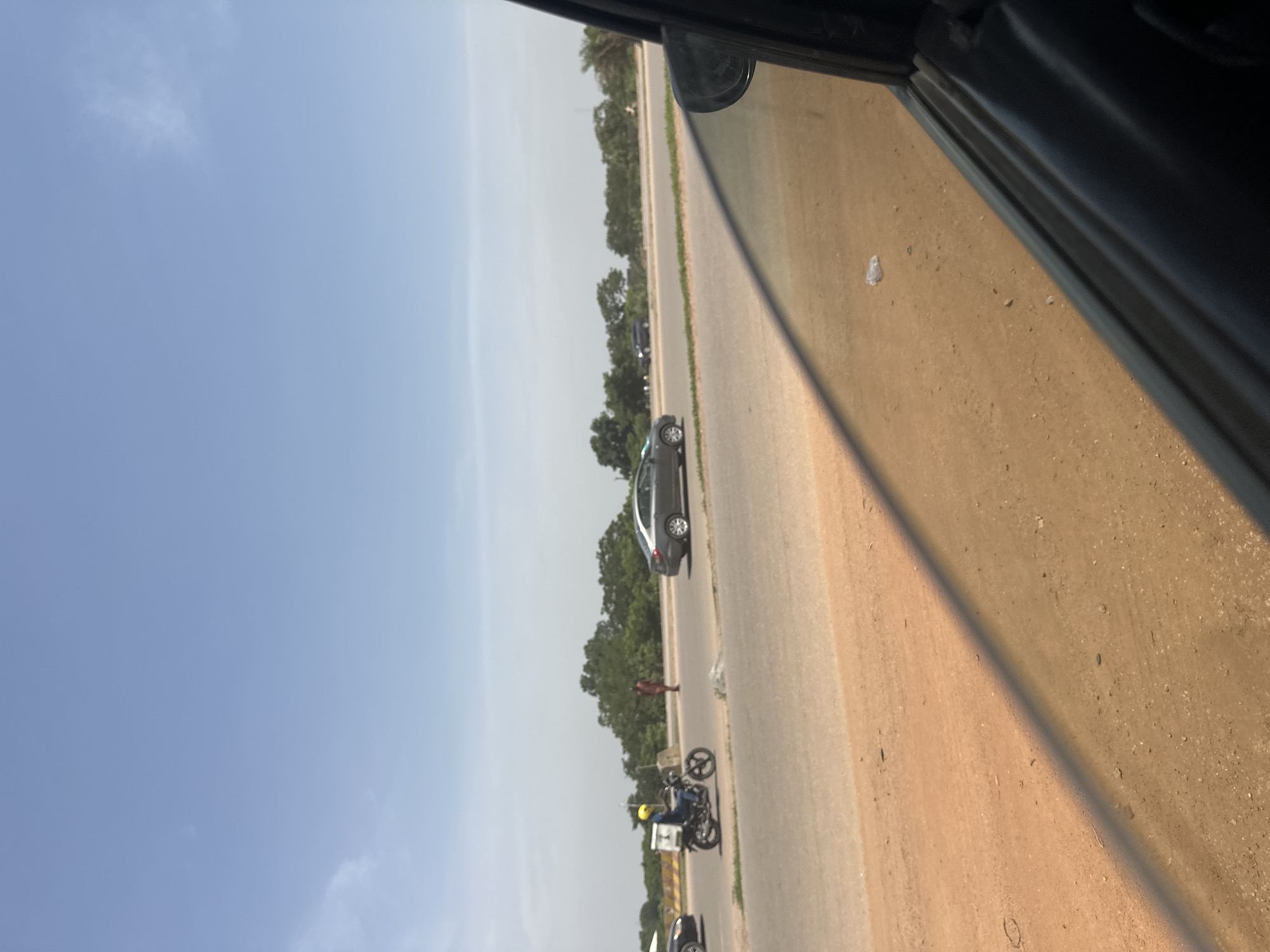 A daytime, eye-level shot captures a side view of a road in Abuja, Nigeria. The road is paved, with a dirt shoulder on the left. A motorcycle carrying a rider with a yellow helmet and a white cooler box travels in the left lane. A pedestrian in a red garment stands on the side of the road. A gray sedan car is in the right lane. Trees line the background. The foreground shows the reflection of a car window and the dirt road. The sky is clear and blue.