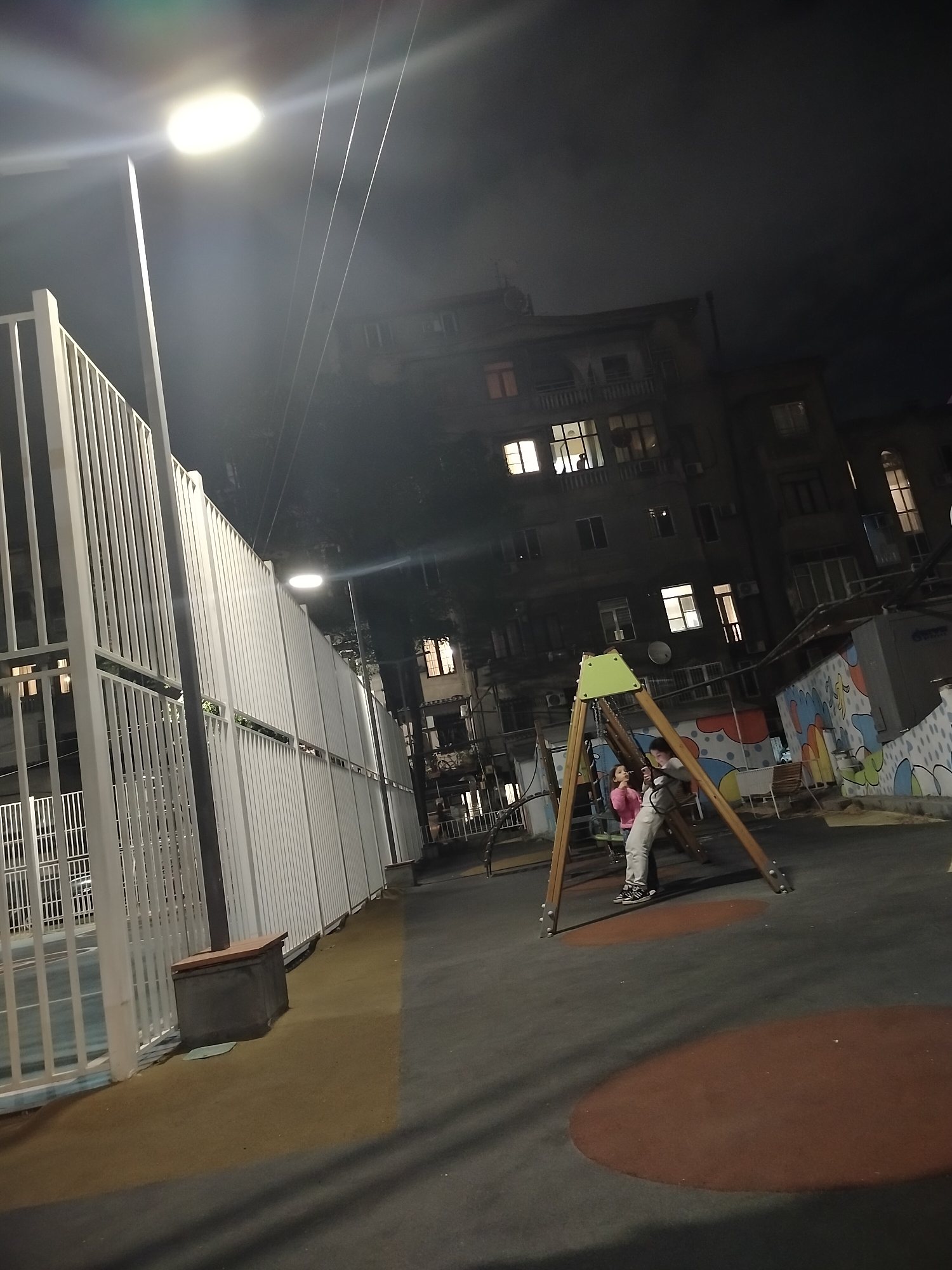 A playground in Yerevan, Armenia at night. Two individuals stand beneath a swing set. A high fence runs along one side of the playground. Buildings stand in the background, some with illuminated windows. A streetlight shines brightly near the fence. The playground surface is a dark material with red circles.