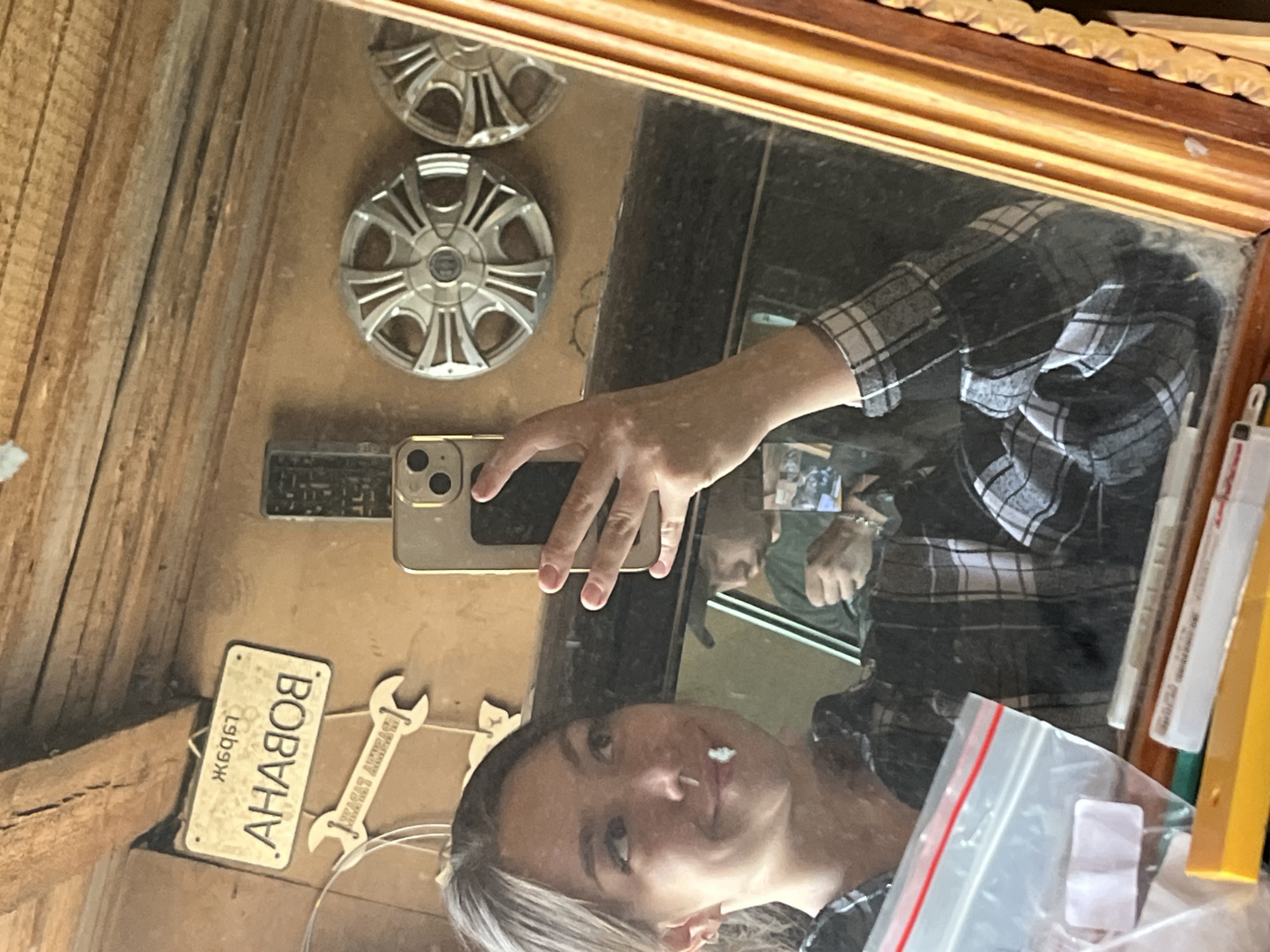 A young woman with brown hair tied back is taking a selfie in a mirror. She is wearing a black and white plaid shirt. There is a white object, possibly cotton, in the corner of her mouth. In the reflection of the mirror, a person wearing a dark t-shirt with a baseball cap is visible. The background of the mirror shows a wall with various objects hanging on it, including a license plate with Cyrillic text, a wrench-shaped decoration, two car hubcaps, and a remote control. The environment appears to be a rustic indoor space, possibly a garage or workshop, with wooden beams visible in the upper part of the image and a wooden frame around the mirror. The overall scene suggests a casual, informal setting.