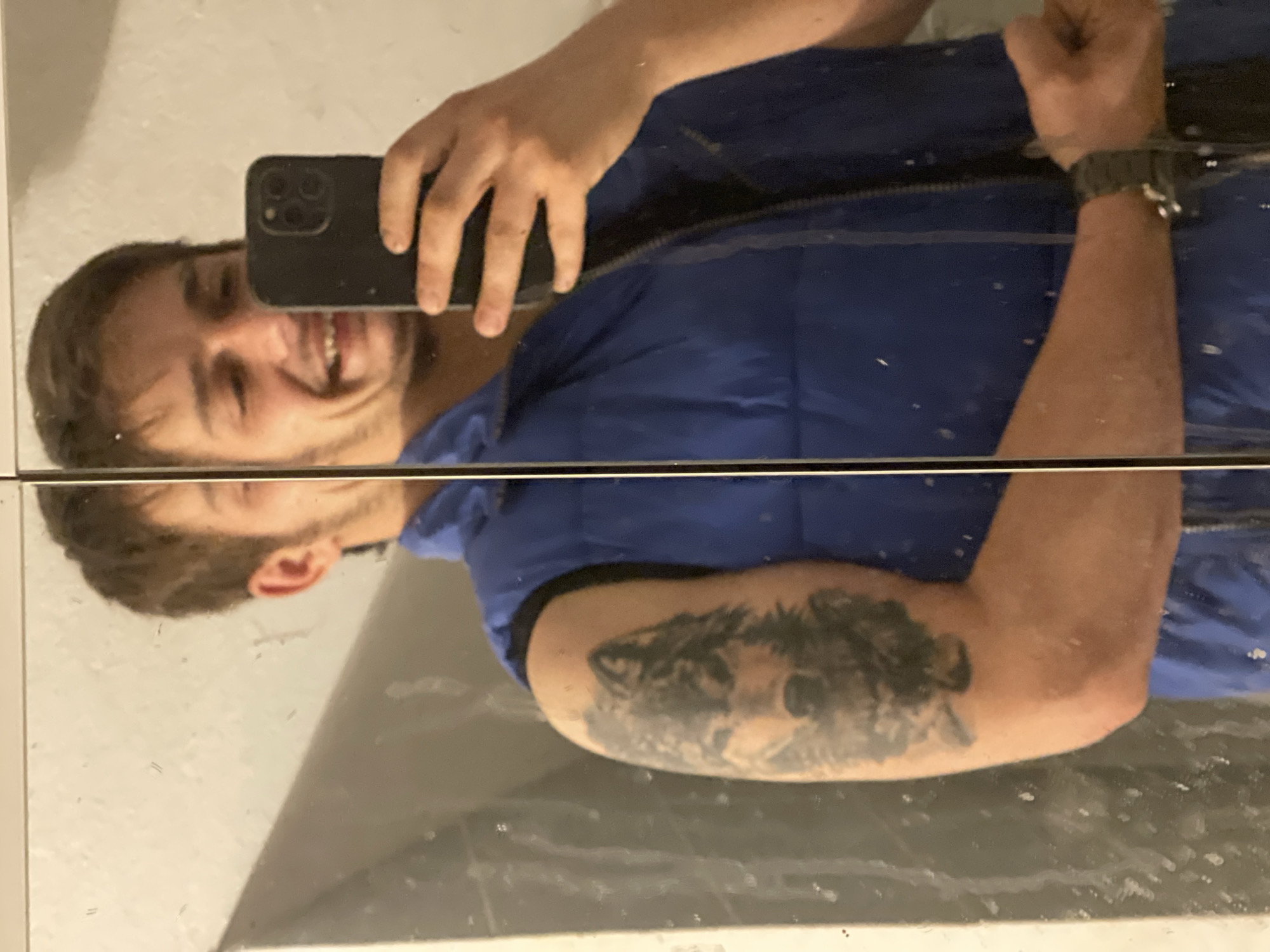 A man is taking a mirror selfie. He is wearing a blue vest and has a tattoo on his left bicep. He is smiling and holding his cell phone. The location is Unknown, Unknown.