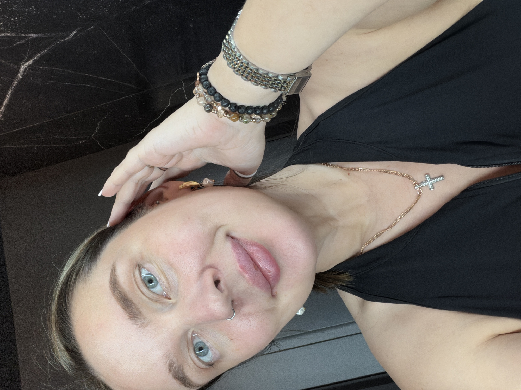 A medium close-up, eye-level shot depicts a woman indoors. She is wearing a black tank top and a gold necklace with a cross pendant. Her left hand is raised to her forehead. She has light skin, light eyes, and a nose ring. She wears multiple bracelets on her left wrist, including one that appears to be a watch. The background is dark with marble-like patterns. The location is not apparent from the image, and it is not possible to determine if it is in Moscow, Russia.