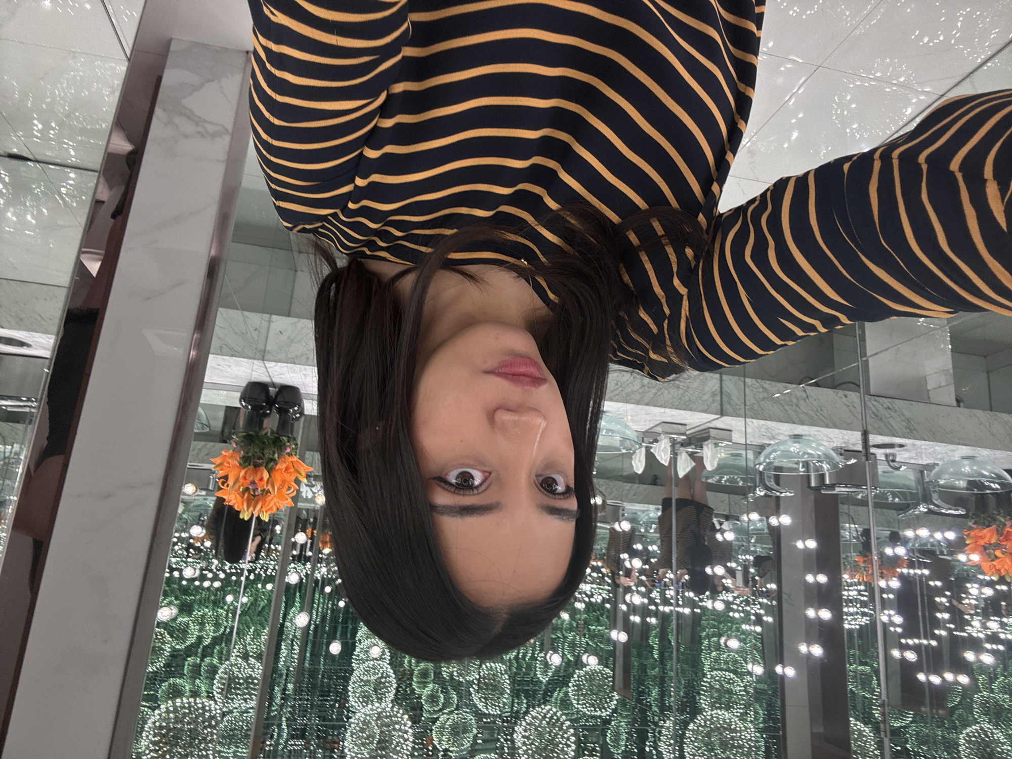 A woman with long, dark hair and wearing a striped shirt (dark with yellow stripes) is holding her phone to take a picture of herself in what appears to be a mirror room. The room is filled with repeating patterns of small, round, illuminated orbs, and there are also flowers and what appear to be hair dryers reflected in the mirrors. Based on the room's appearance and decorative elements, it is potentially an art installation, though this cannot be confirmed from the image alone. The location of the photograph is in Puebla, Mexico.