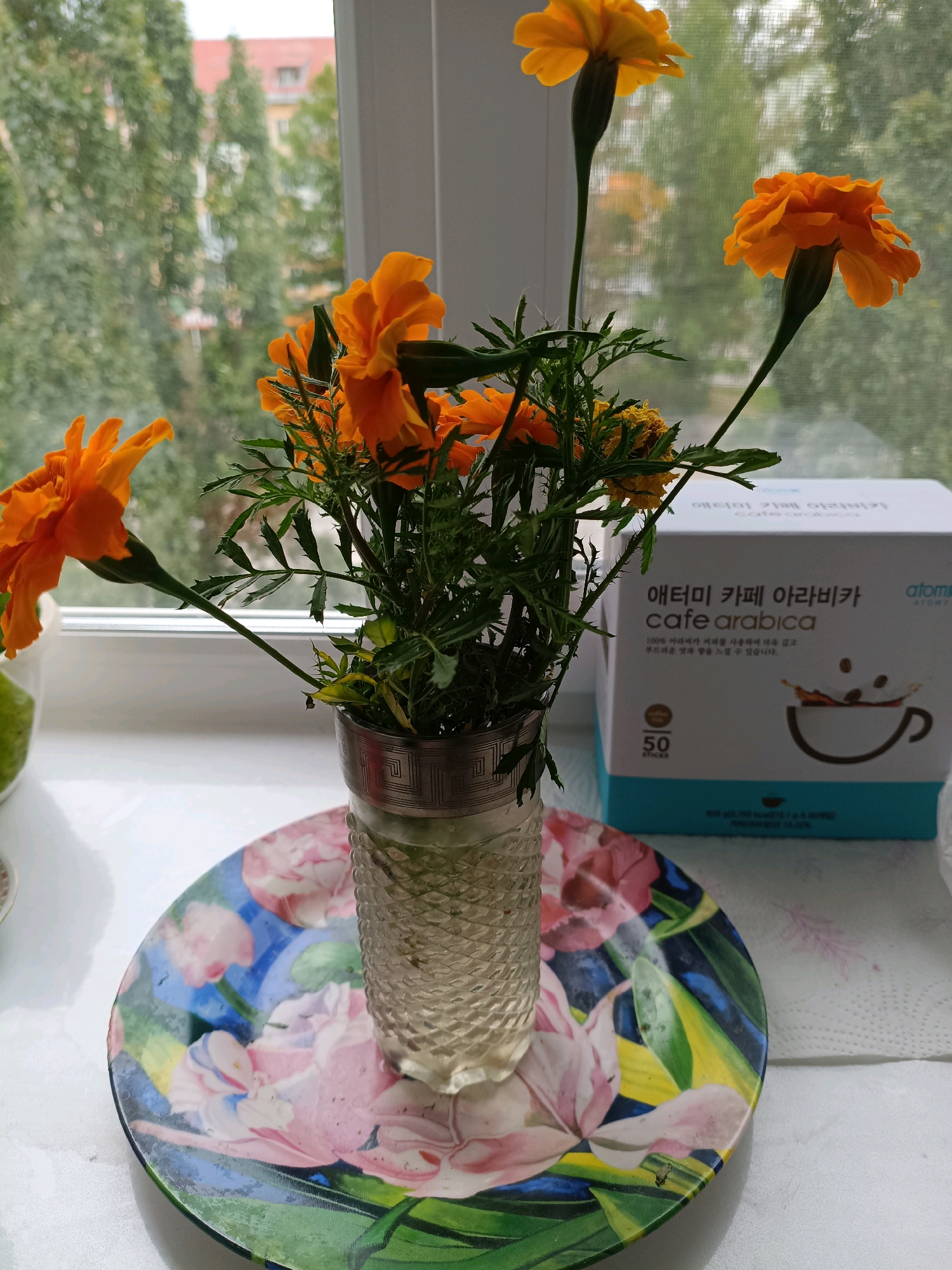 A still life arrangement sits on a windowsill, featuring a bouquet of bright orange marigolds in a textured glass vase. The vase rests on a vibrantly decorated plate with floral motifs in pink, blue, yellow, and green. Behind the flowers, a box of "Atomy Cafe Arabica" coffee sticks is visible, with Korean text and an illustration of coffee splashing into a cup. The background shows a blurred view of trees and buildings through the window, suggesting an urban or suburban setting. The image was taken on a windowsill in Asbest, Russia.
