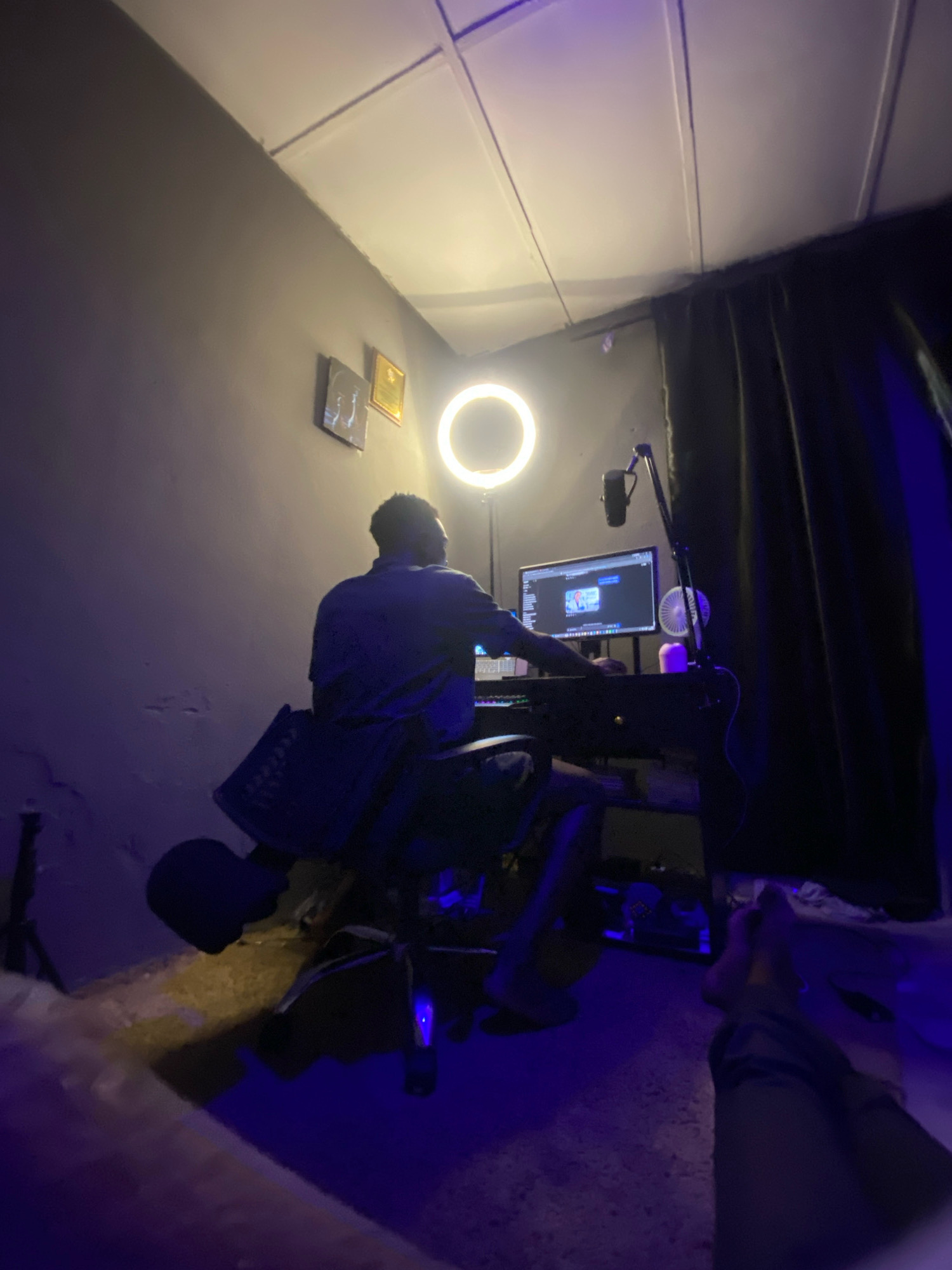 The image depicts an indoor scene, possibly a home studio, illuminated by a ring light and blue ambient lighting. A person is seated in a chair facing a computer setup, which includes a monitor displaying content, a keyboard, and a microphone on an articulated arm. The room has a dark-colored wall with two framed pictures hung on it, and a black curtain covers a portion of the right side of the room. The ceiling appears to be made of acoustic tiles. The floor is partially visible, covered by a light-colored rug. The overall atmosphere suggests a creative or work-related activity. No specific details about the city of Jos, Nigeria, are discernible from the image itself.