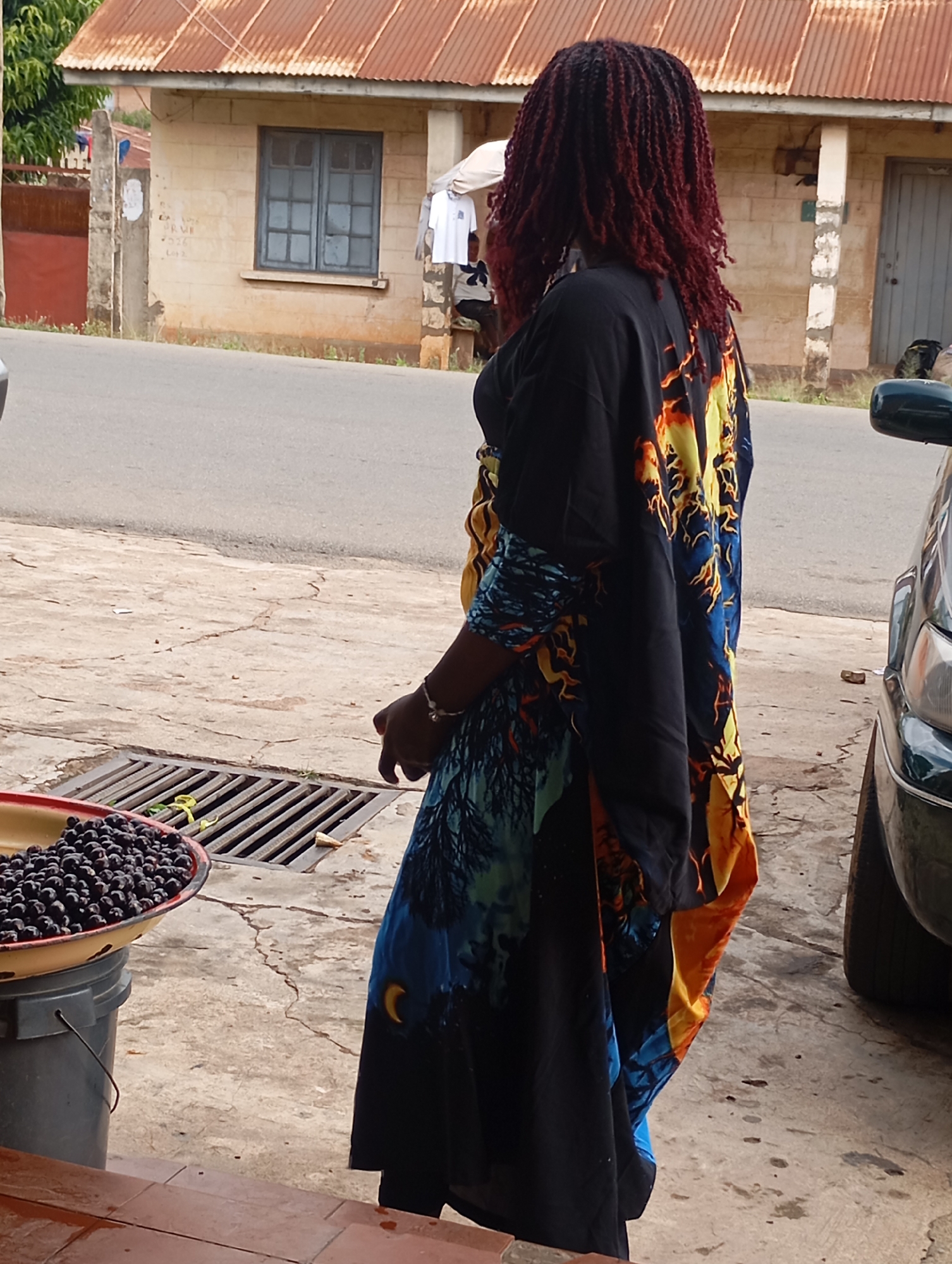 A Black woman with dark, red-brown braided hair, wearing a black garment with a colorful flame and moon pattern, stands facing away from the camera on a concrete surface in front of a building. To her left, a shallow, yellow-rimmed bowl sits atop a gray bucket, filled with dark, small, round fruits, likely berries or plums. A metal grate with yellow ties lies on the ground behind the bowl. The building behind the woman has a corrugated metal roof and pale brick walls. A blue-paned window is visible on the building. A dark-colored car is parked to the right of the woman. The scene appears to be outdoors, on a street or sidewalk in the city of Jos, Nigeria.