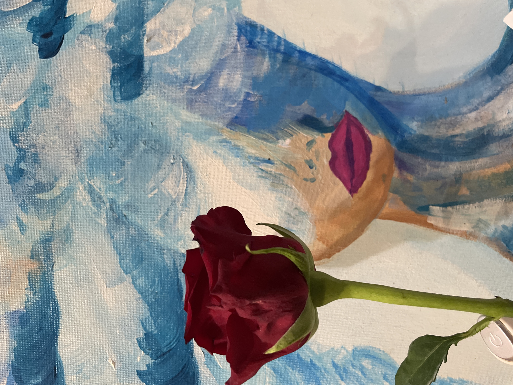 A close-up shot reveals a dark red rose bud laid against a painted canvas. The canvas displays a figure executed in blue and white brushstrokes with an area of tan skin-toned paint that has a distinct reddish-purple element resembling lips. The rose is fully closed; the stem is green with leaves emerging from the stem. A small, white power button is attached to the stem. The canvas texture is visible. The setting is in the city of Sochi, Russia.