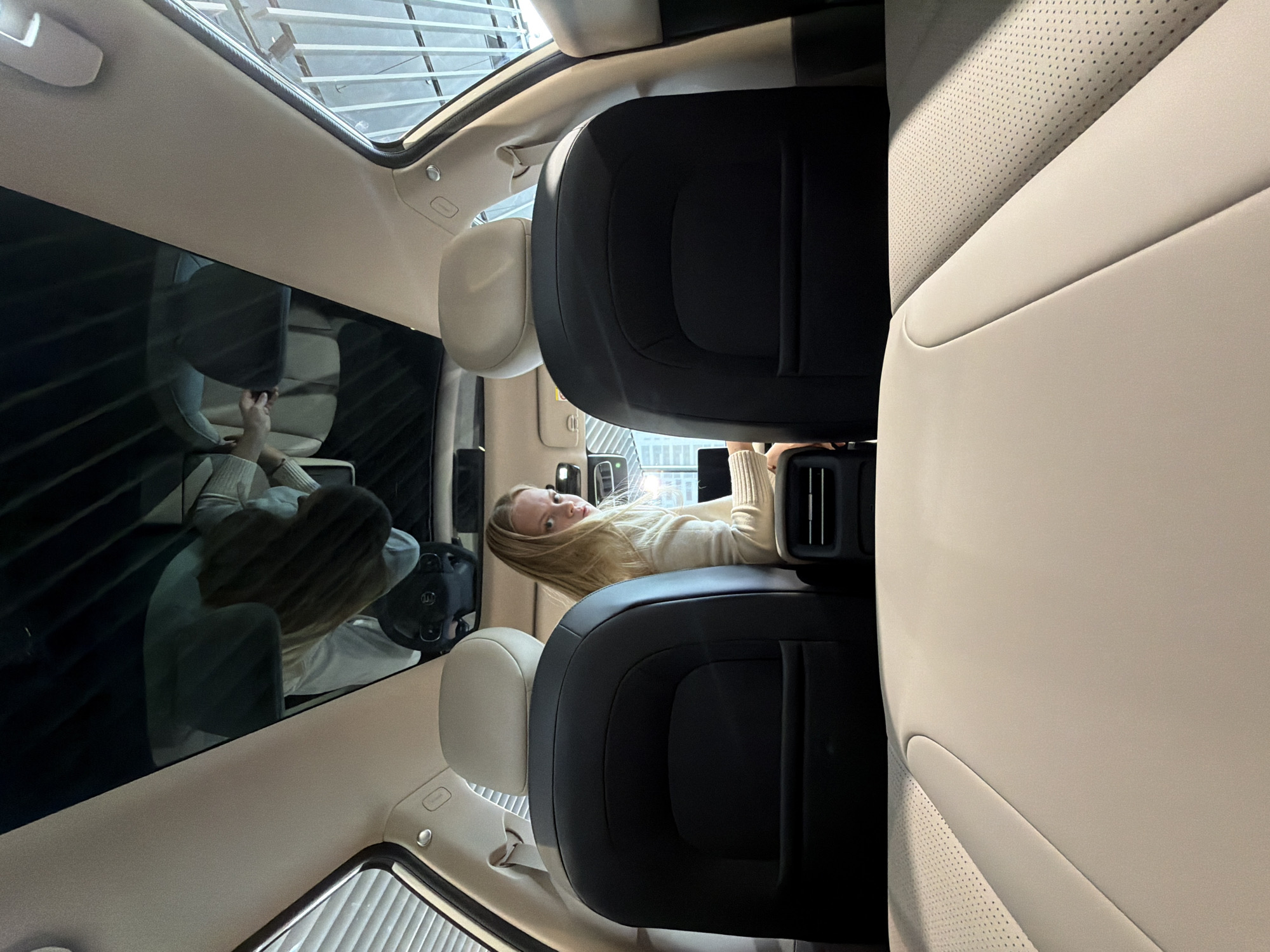 A woman with blonde hair is in the back seat of a car, holding a phone. She is wearing a light-colored sweater. The car's interior features black leather rear seats and lighter colored leather front seats. A driver is visible in the front, reflected in the car's glass roof. The setting appears to be indoors, under a glass structure, which could be a showroom or similar environment. The city, Izhevsk, Russia, is not visually verifiable from the image.
