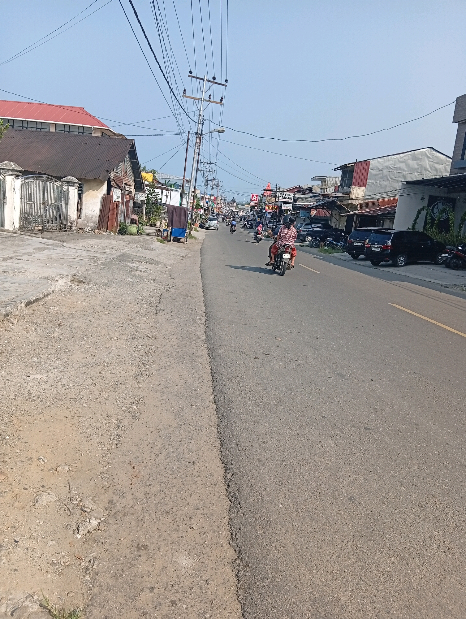 The image captures a street scene in the City of Gunungsitoli, Indonesia, under a clear, bright sky. The road, asphalted with a yellow dividing line, is flanked by a paved sidewalk on the left and a row of buildings on the right. The buildings, some appearing aged with corrugated metal roofs and weathered facades, stand adjacent to newer structures with white and red accents. Power lines crisscross the sky above.
On the left side of the road, a dilapidated building with a rusty corrugated roof is visible, next to a white gate. Further down the street, a more modern building with a red roof is seen. The right side of the road features various commercial establishments, including what appears to be an optician ("OPTIK MATAHARI") and a store labeled "GLOBAL". Several parked vehicles, including black SUVs and motorcycles, line the street.
Traffic is present, with a man and a woman riding a motorcycle in the foreground, moving away from the camera. Other motorcycles and a few cars are visible further down the street, indicating moderate activity. A person can be seen standing near a cart on the left side of the road. The ground on the left is a mixture of rough pavement and dirt. The overall impression is that of a busy, developing urban environment.