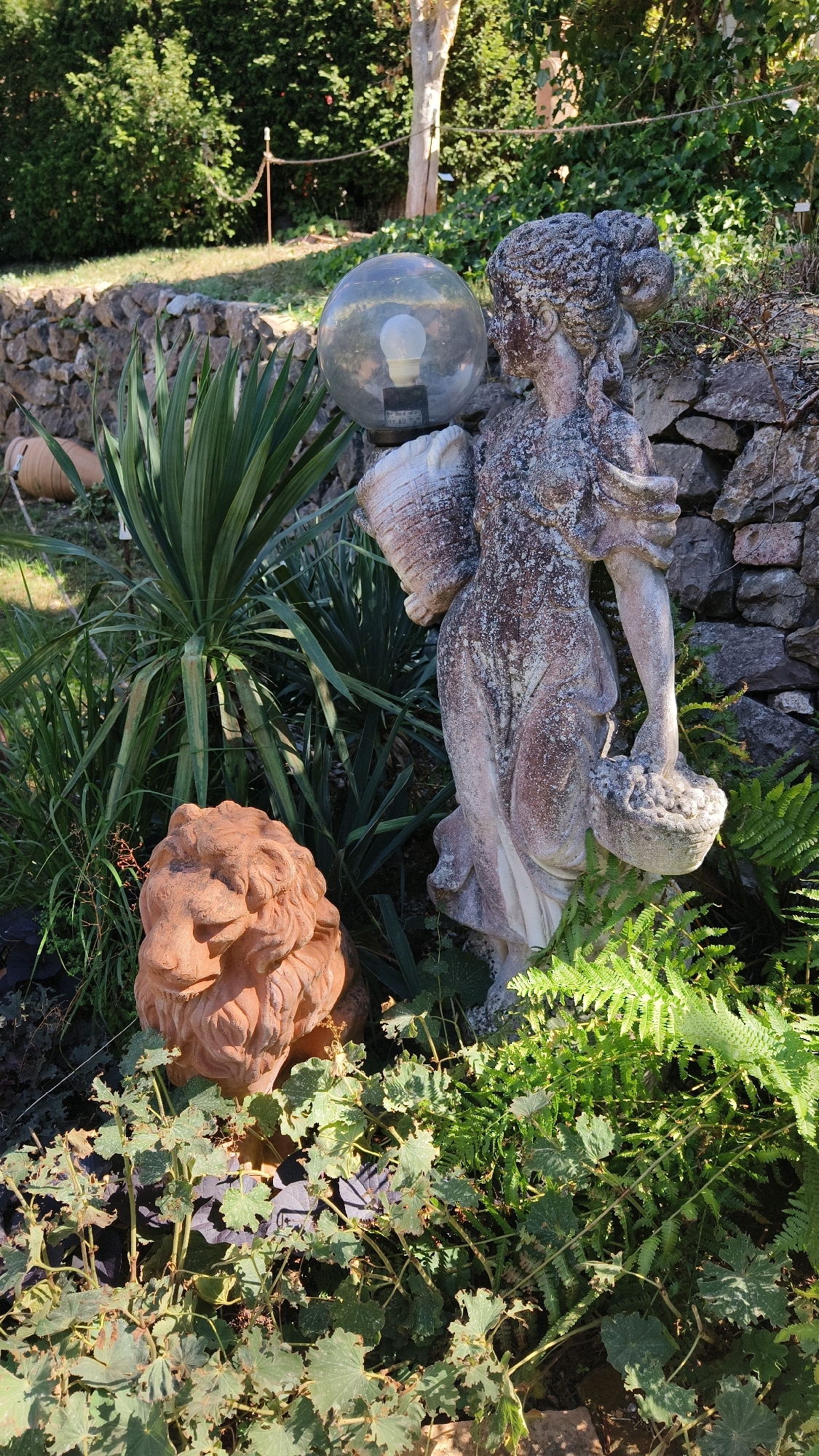 The image depicts a garden scene with several decorative items. In the foreground, a terracotta lion's head statue is visible, partially obscured by green foliage. To its right, a weathered statue of a woman holding a basket and a globe lantern stands among ferns and other plants. The background features a dry stone wall, a yucca plant, and lush green trees and bushes. A rope barrier with wooden posts is also present in the background, suggesting a demarcated area. The lighting suggests it is daytime, with sunlight casting shadows on the scene. The location is specified as Langenlois, Austria.