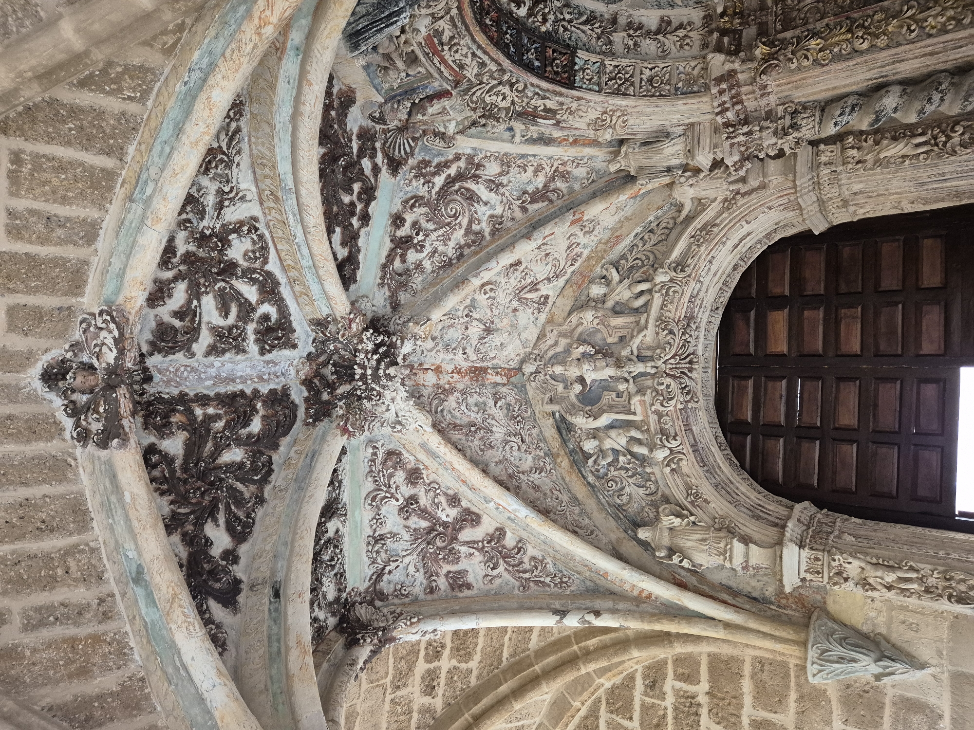 The image shows an ornate architectural detail of a building's interior, likely a church or a historical monument. The focal point is a vaulted ceiling or archway adorned with intricate carvings and painted decorations. The architectural style appears to be Gothic or Renaissance, characterized by elaborate ornamentation and the use of stone and plaster.
The vaulting is composed of ribbed arches, with each section filled with detailed plasterwork. These decorations feature floral motifs, swirling patterns, and possibly cherubic figures or religious iconography. The colors used are muted, with shades of white, beige, brown, and subtle hints of blue and green, suggesting age and weathering.
On the sides, rough-hewn stone blocks form part of the structure, contrasting with the refined decoration of the interior. Below the archway, a large, dark wooden door is visible, indicating an entrance. The lighting is somewhat dim, casting shadows that accentuate the three-dimensional qualities of the carvings.
The image is taken from a low angle, looking upwards at the ceiling and the arched entrance. The overall impression is one of historical grandeur and artistic craftsmanship.