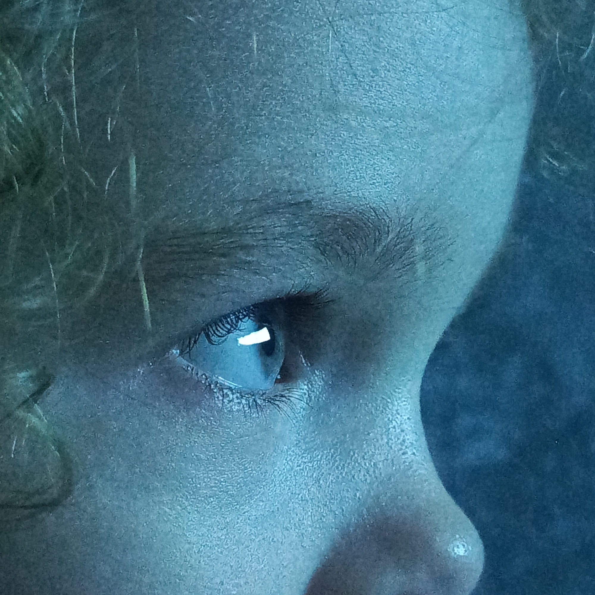 The image is a close-up shot of a person's eye, predominantly illuminated with a blue-green hue. The eye is open, with eyelashes visible, and a bright reflection is present in the pupil. The skin surrounding the eye appears textured, with fine lines and pores discernible. The background is dark and blurred, with some lighter streaks that could suggest movement or a textured surface. The overall impression is one of intense focus or perhaps a digital glow. There is no discernible context that would allow for determining the location in Марковский, Russia, or any specific setting.