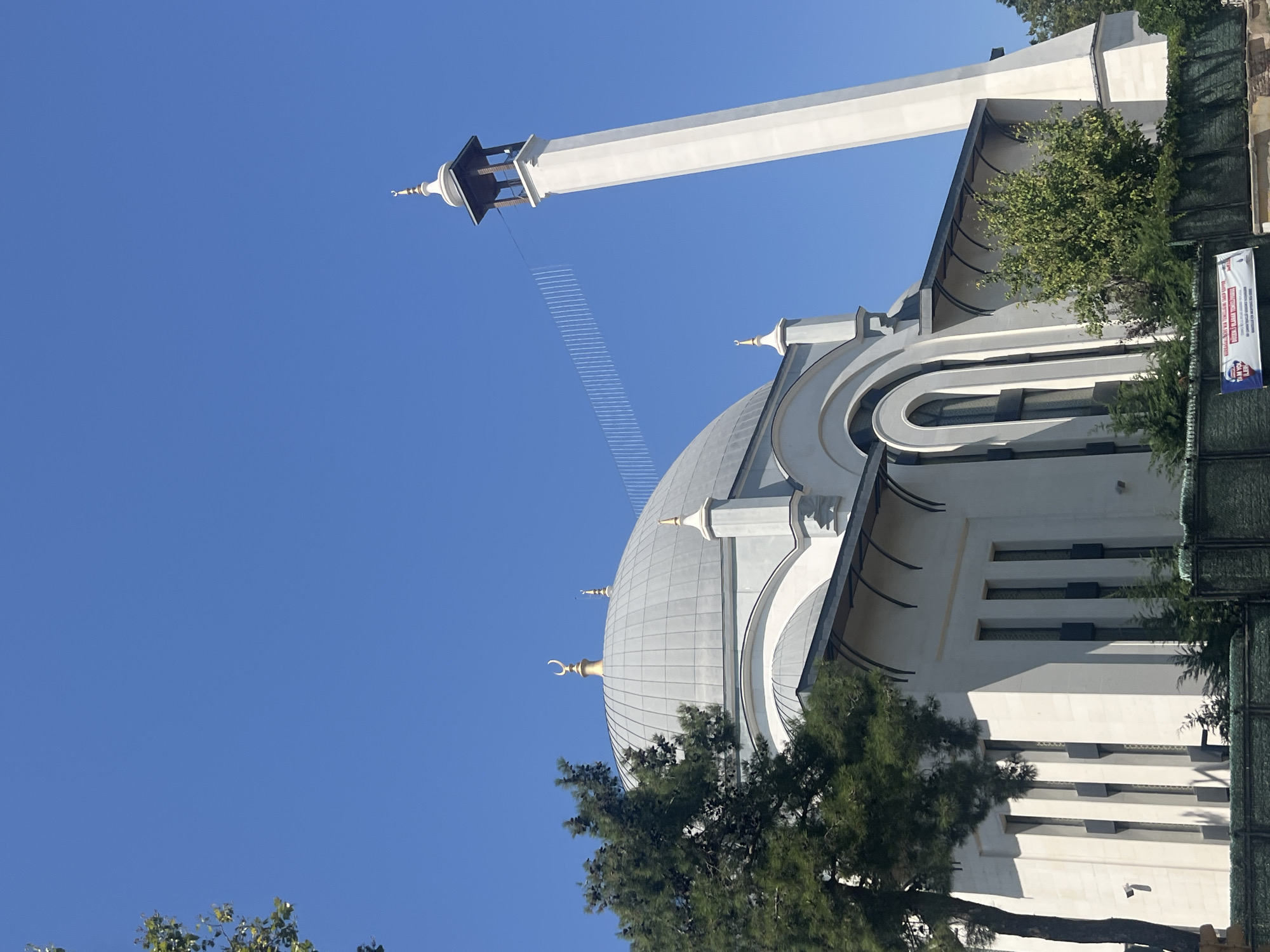 A low-angle, full-shot photograph captures the exterior of a mosque in Kadıköy, Turkey, under a clear, bright blue sky. The mosque features a prominent white minaret topped with a golden crescent and finial, and a large gray dome adorned with a similar golden crescent. The building's facade is white with arched windows and decorative elements. A dense green pine tree stands to the left of the mosque, partially obscuring the dome. In the foreground, a dark green fence runs horizontally across the image, with some signage visible at the bottom right. The overall scene depicts a sunny day in an urban environment, highlighting the architectural beauty of the mosque.
