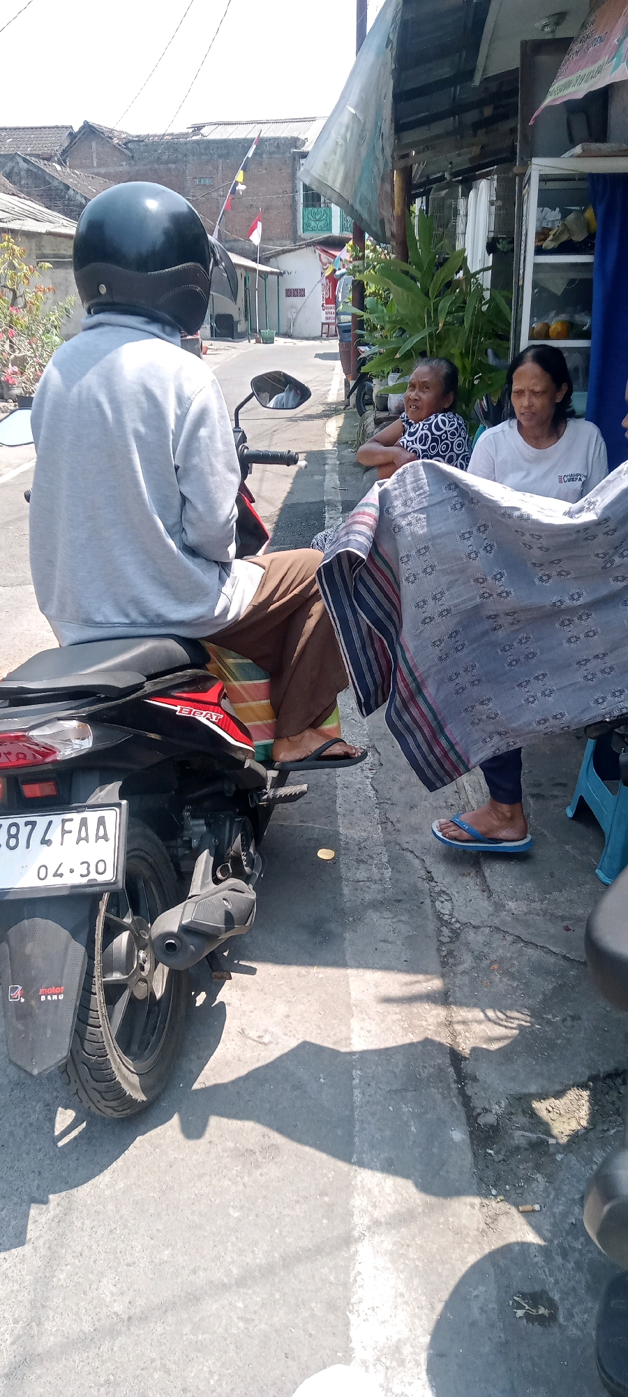 A person wearing a gray hooded sweatshirt and brown pants sits on a black motorcycle. They are wearing a black helmet. The motorcycle has a license plate that reads "B 874 FAA 04.30". To the right of the motorcycle, two women are seated. The woman closer to the motorcycle is wearing a black and white patterned shirt. The woman to her right is wearing a white t-shirt. A piece of patterned fabric, possibly a sarong, is draped over the lap of the woman in the white t-shirt and extends towards the motorcycle. In the background, there are buildings with tiled roofs and some greenery. The scene appears to be on a street in Surakarta, Indonesia, with the architecture and the motorcycle suggesting a local context. The ground is paved and there are shadows cast from the overhead structures and objects.