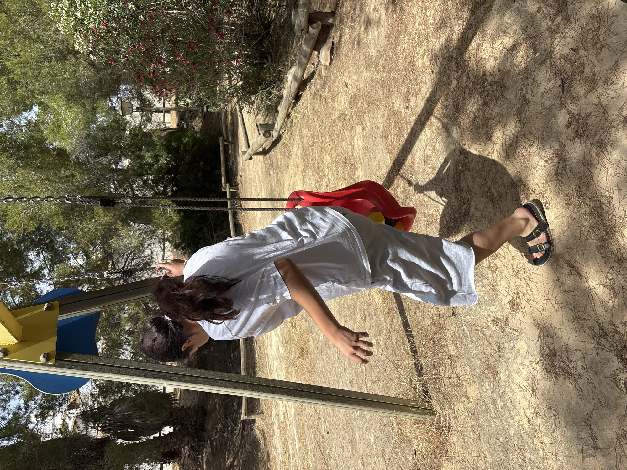 A young girl with dark hair is swinging on a playground swing. She is wearing a white t-shirt and light-colored pants. The swing set has a yellow and blue structure. The swing seat is red. The ground is covered in sand and dry pine needles. In the background, there are trees and some pink flowers. The sun is casting shadows, suggesting it is daytime. The overall setting appears to be a park or playground. The girl's posture and outstretched arm suggest she is in motion, likely enjoying the swing. There is no visible text in the image. The location context provided is Orihuela, Spain.