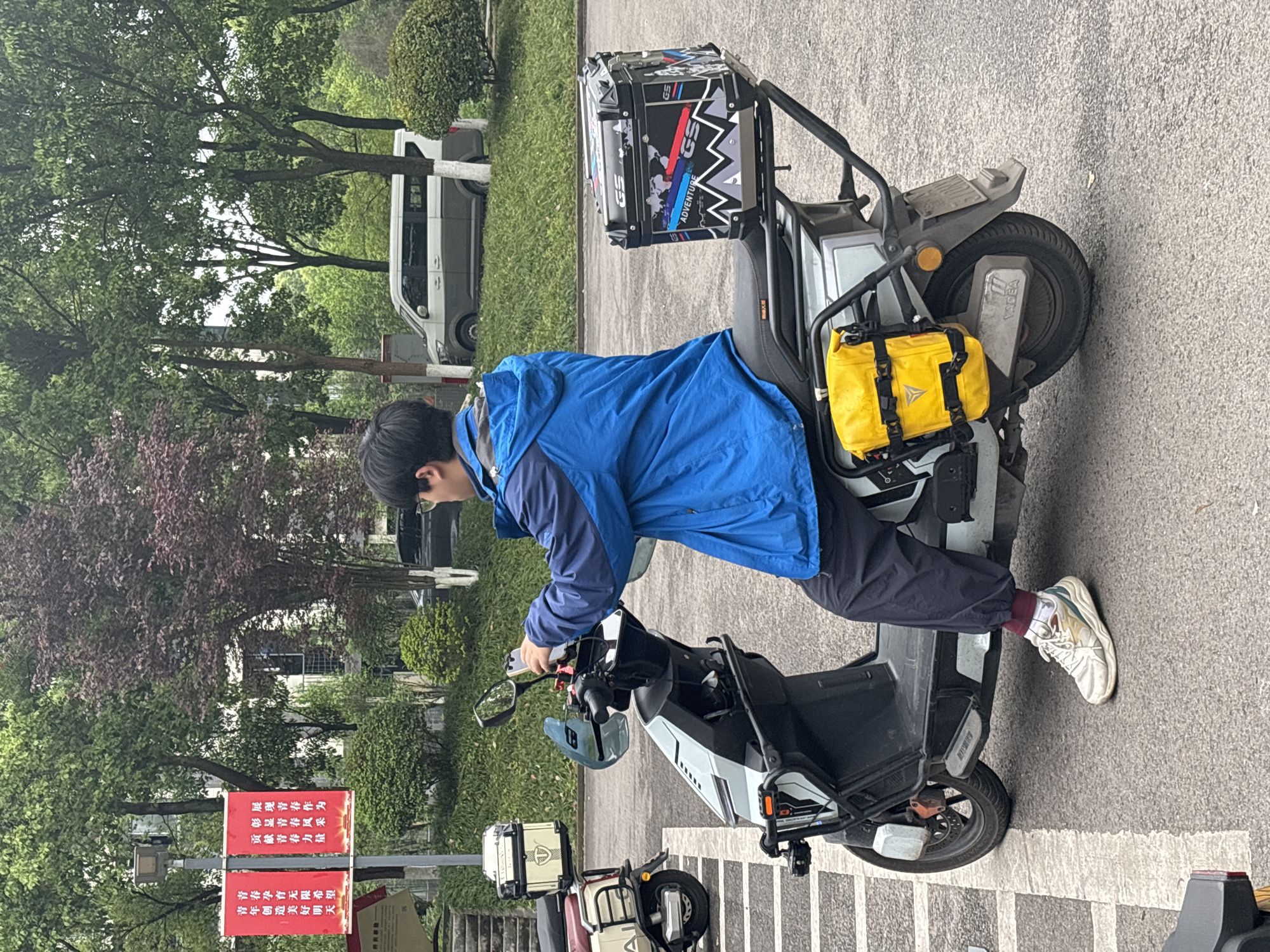 A man in a blue jacket and black pants is operating a Yadea electric motorcycle on a paved area. The motorcycle is equipped with a top case labeled "GS Adventure" and a yellow side bag. A sign with red Chinese characters is visible. The setting appears to be in Jiangjin, China. A white van is parked in the background.
