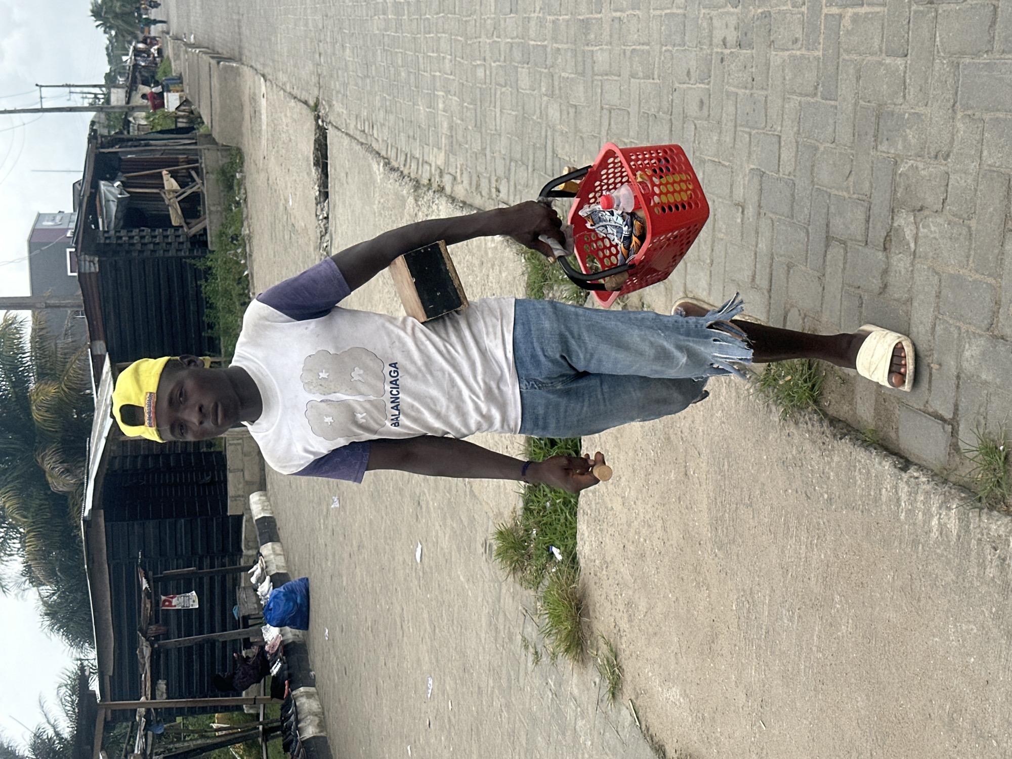 A young Black male wearing a yellow baseball cap, a white t-shirt with purple sleeves, and distressed denim shorts walks along a paved street in Badagry, Nigeria. He carries a wooden box under his right arm and a red plastic basket filled with goods in his left hand. He holds a small, round object in his right hand. The street is lined with small, open-air market stalls and buildings made of corrugated metal and wood. In the background, palm trees are visible, and several people can be seen at the market. The ground is littered with some debris. The sky is overcast.