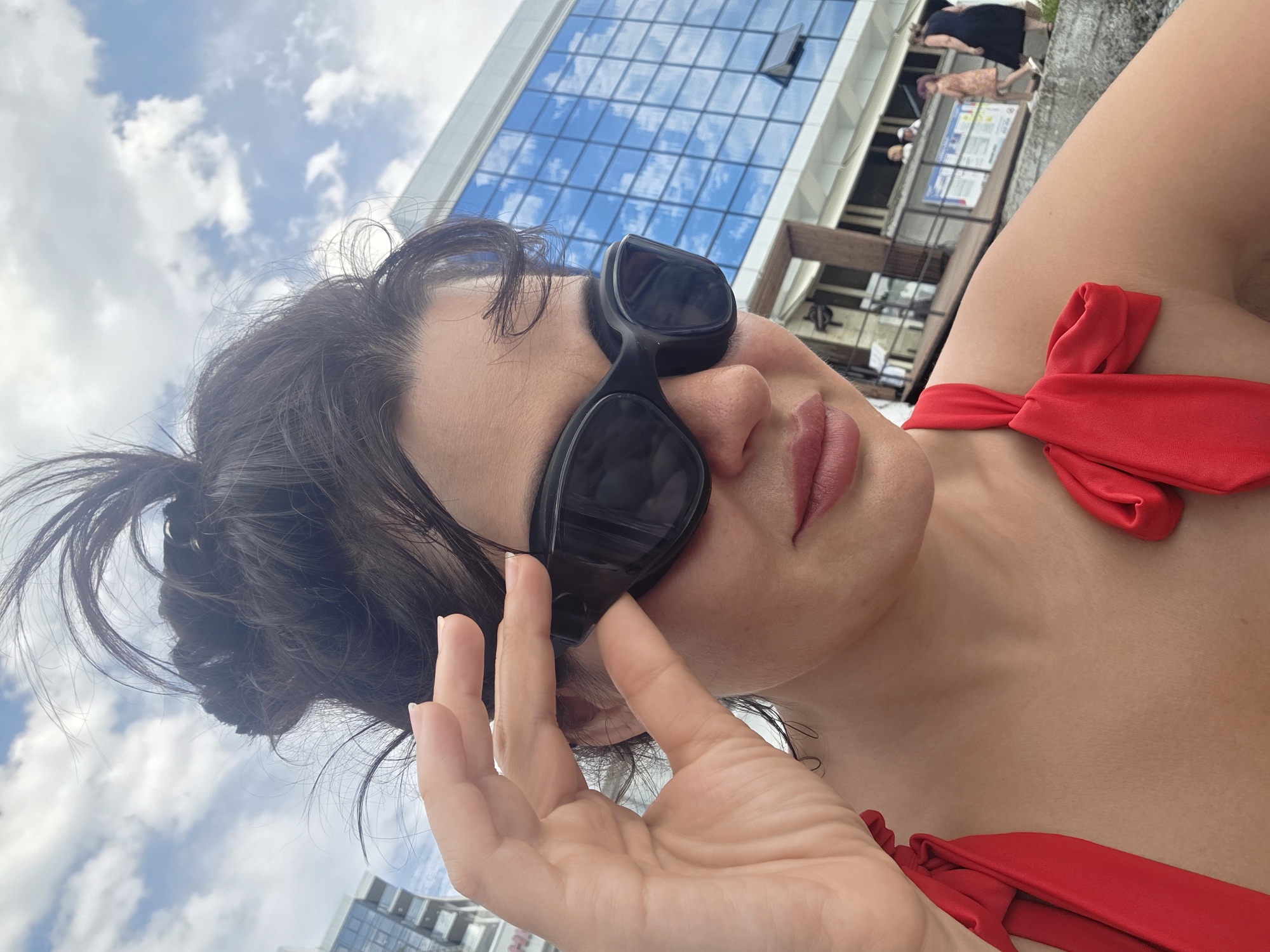 A woman with dark hair pulled up in a messy bun is wearing large, black sunglasses and a red swimsuit. She is holding her sunglasses on with her right hand. The background shows a modern building with blue glass windows, a cloudy sky, and other people walking in the distance. The scene appears to be outdoors in a city, possibly a coastal area, given the swimwear and the building's architecture. This location is consistent with the seaside resorts and modern architecture found in Sochi, Russia.
