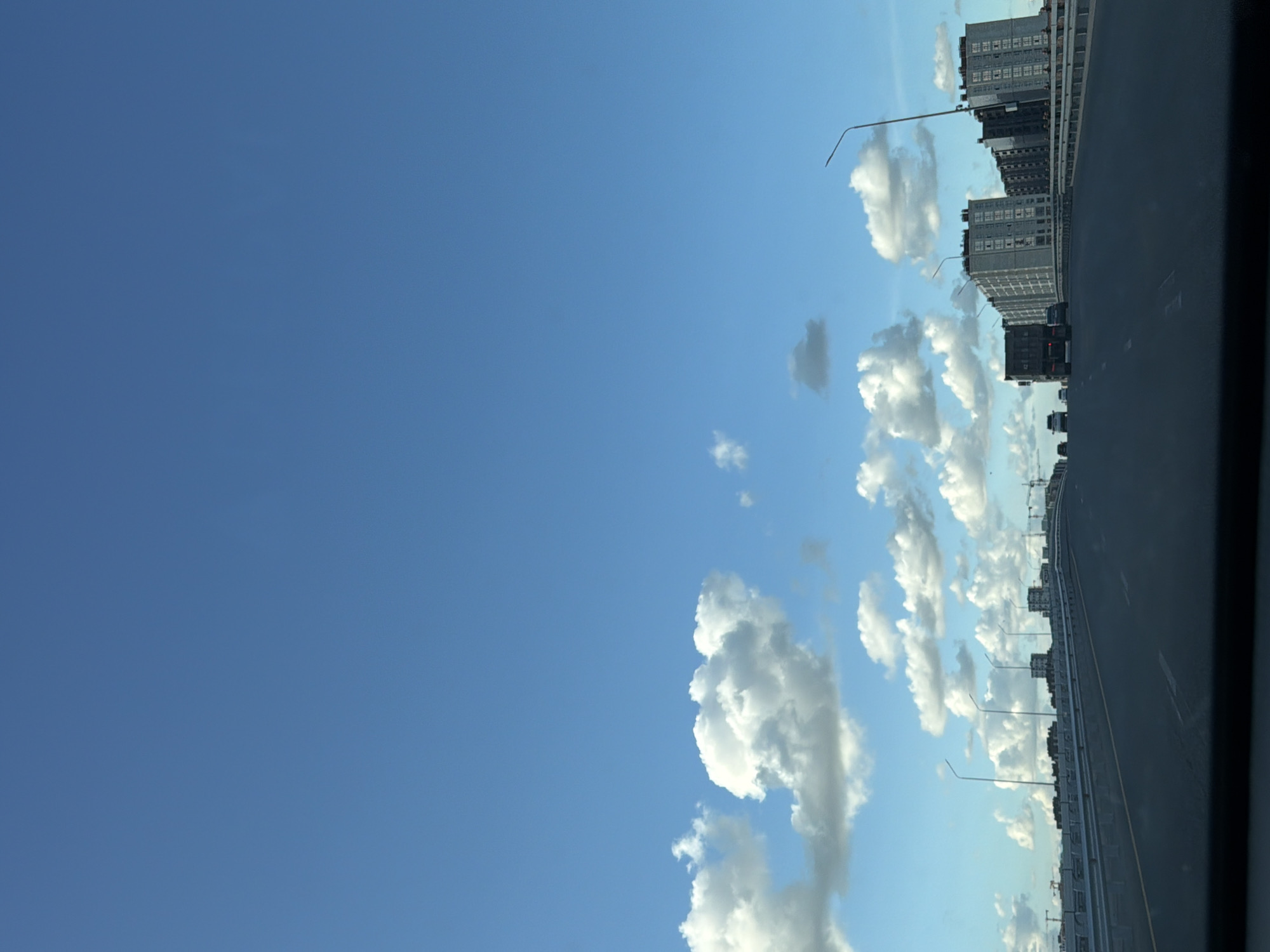 The image depicts a view from a vehicle on a multi-lane highway or bridge, likely in Astana, Kazakhstan, given the architectural style of the buildings and the presence of construction cranes. The sky is a clear, bright blue with scattered white, fluffy cumulus clouds, some of which are casting shadows. A prominent feature is a series of modern, high-rise apartment buildings with repeating window patterns and balconies on the right side of the frame. On the left, a long, low barrier with railing lines the edge of the roadway, and several streetlights are visible. Several vehicles are present on the road ahead, including a large truck in the center lane, and passenger cars. In the distance, construction cranes are visible above the buildings, indicating ongoing development. The overall scene suggests a contemporary urban environment under a pleasant, sunny sky.