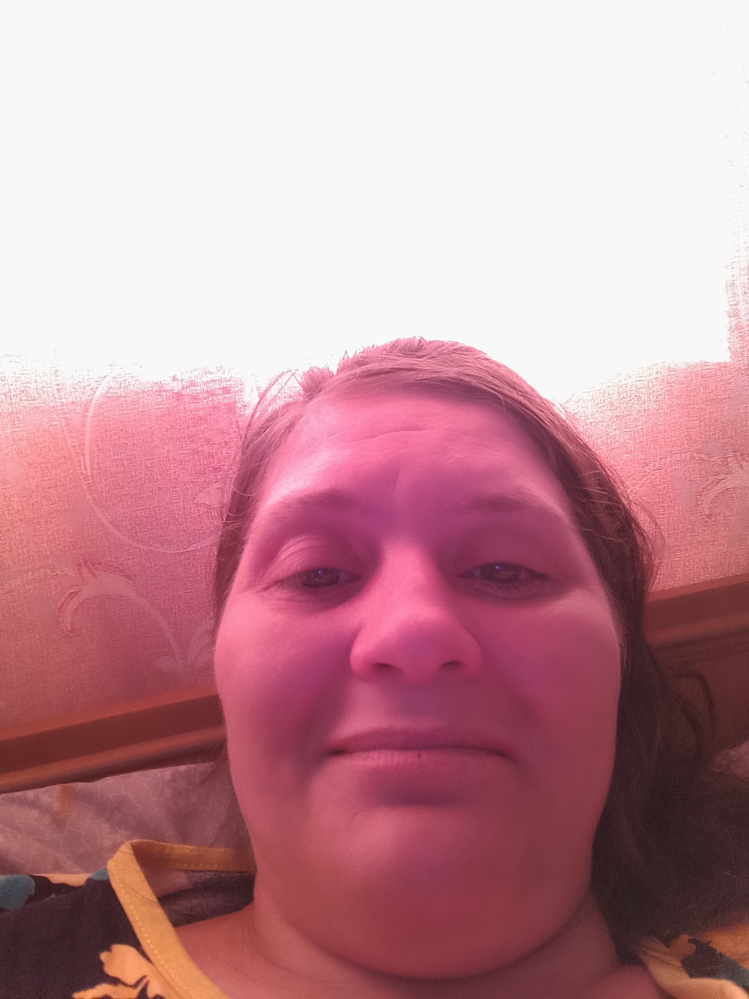 A close-up, low-angle, selfie-style photograph shows a woman's face. She has fair skin, brown hair, and is looking directly at the camera. Her eyes are open and her lips are slightly parted. The lighting in the image is predominantly pink, casting a warm glow on her face and the textured wallpaper behind her. The wallpaper has a subtle, patterned design. The woman is wearing a patterned top with a yellow neckline. The scene appears to be indoors, possibly in a bedroom, given the presence of bedding and a headboard visible in the lower right portion of the image. The focus is on the woman's face and upper body.