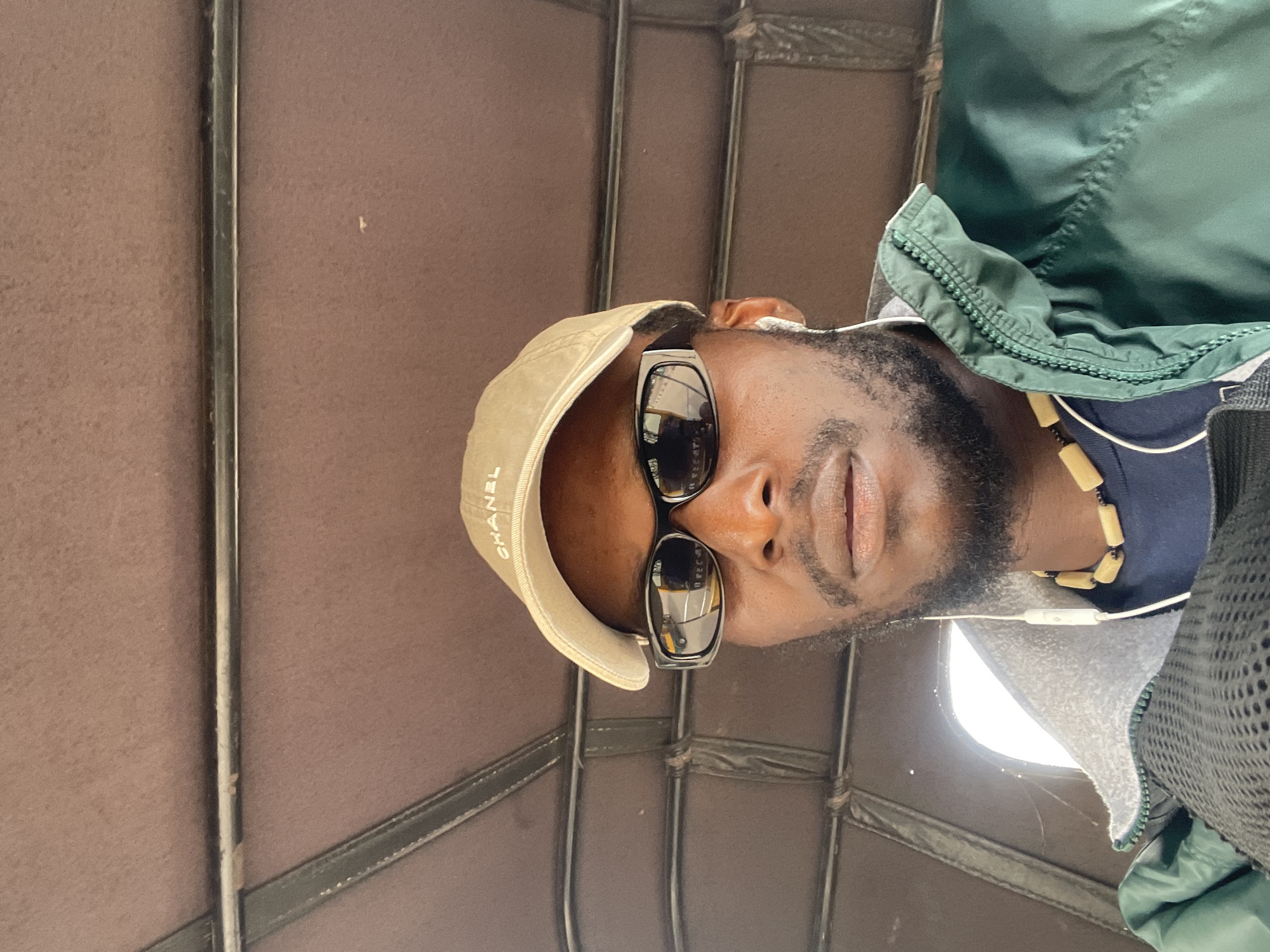 A medium shot captures a Black male wearing a tan baseball cap with "CHANEL" embroidered on it, black wraparound sunglasses, a green jacket with a zipper partially open, and a beaded necklace. He is wearing white earbuds, and a portion of his navy blue shirt is visible. The subject is looking directly at the camera. The background suggests the interior of a vehicle, possibly a rickshaw, with brown padded lining and metal bars. The scene appears to be a street in Jos, Nigeria, given the context provided.