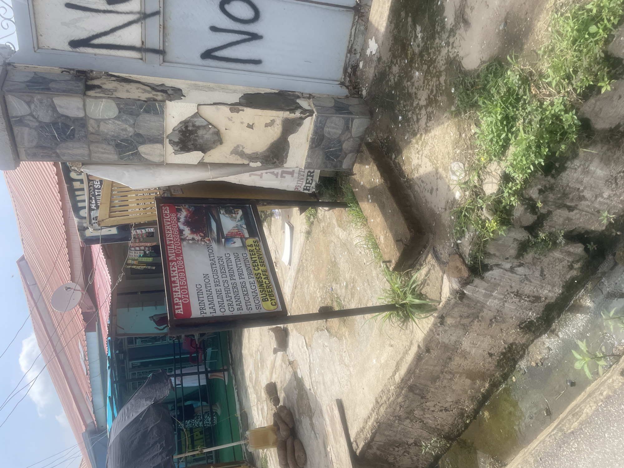 This is a vertical, eye-level, medium shot of a storefront in Abuja, Nigeria. The storefront, identified by visible text as "ALPHALAKES MULTISERVICES", appears to be a business center and cybercafe offering printing, lamination, online registration, graphics design, banners, stickers, and signage printing. The building facade is weathered, with peeling paint and some decorative stone tiles. A sign on a metal pole in front of the store lists the services and contact numbers.
The scene shows a street in front of the establishment, with a concrete drainage channel running alongside. Green weeds are growing in and around the channel, suggesting recent rain or ongoing irrigation. The ground is paved with concrete and dirt. The sky is visible in the upper left quadrant of the image, indicating it is daytime. No people are clearly visible, though there might be figures inside the shop through the glass. The overall impression is of a busy, functional urban environment with some signs of wear and tear. The graffiti on the metal gate above the storefront, including the word "No," adds a layer of urban character to the scene. The image is taken during the day, with natural lighting illuminating the area.