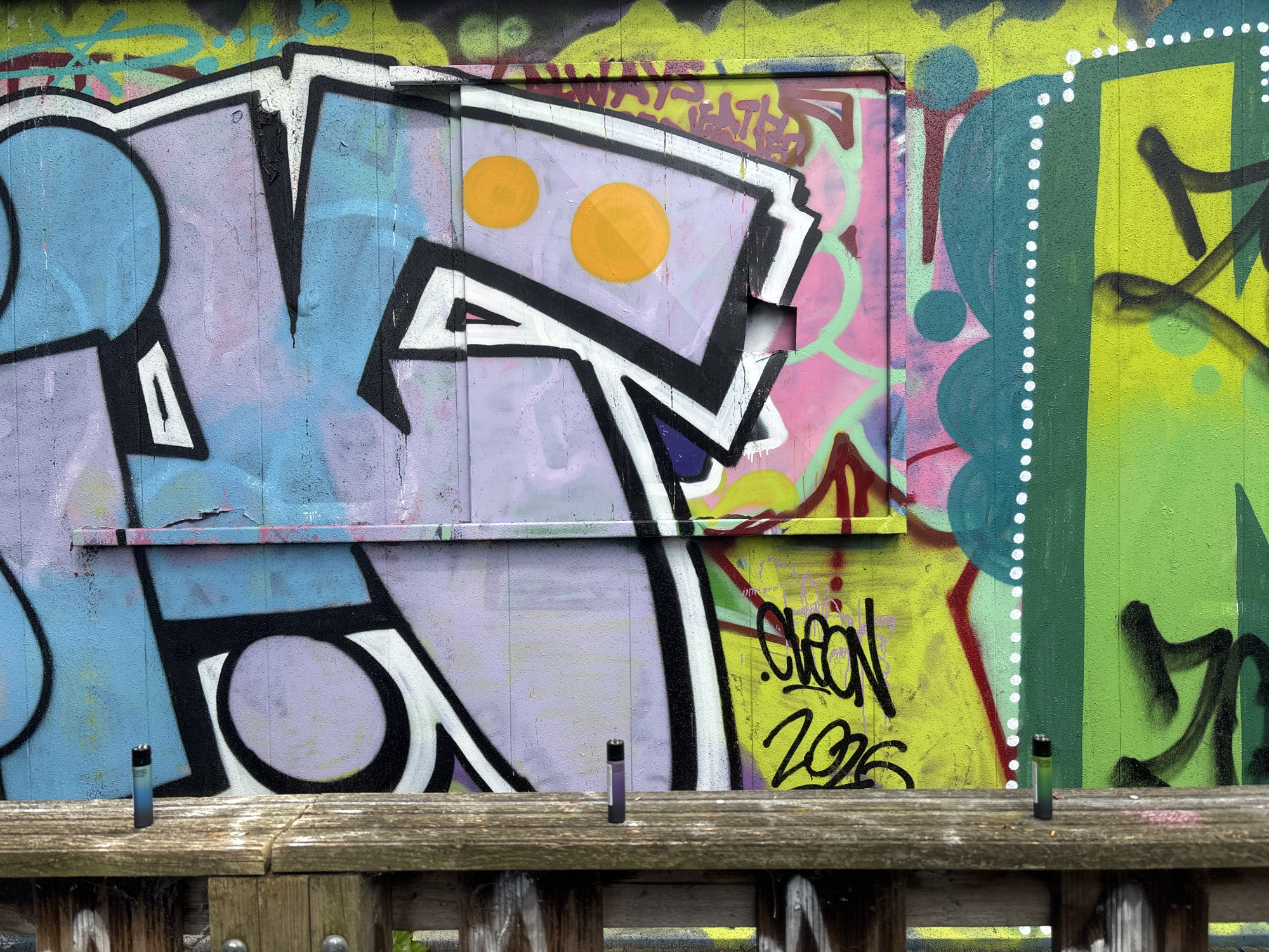 A close-up photograph captures graffiti art on a wooden fence. The graffiti includes letters and abstract shapes in blue, purple, yellow, and green hues, with black outlines. A small rectangular form with two orange circles is visible. The text "CLEON 2026" is also part of the graffiti. The wooden fence in the foreground has vertical slats and horizontal boards, with some metal fasteners. The location appears to be an urban environment, possibly London, United Kingdom, given the style of graffiti and the general urban setting.