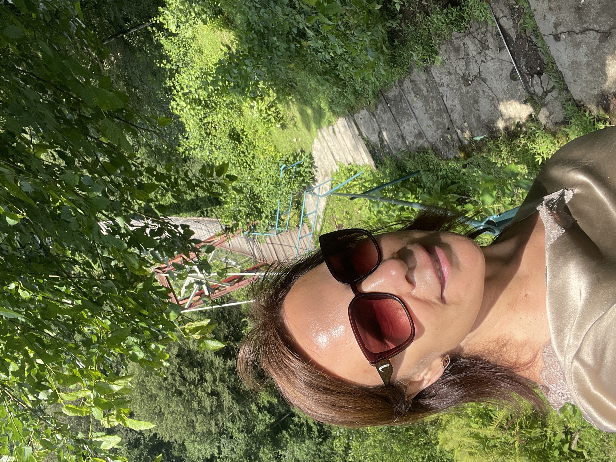 In Tqwarchal, Abkhazia, a woman with brown hair, wearing large sunglasses and a satin top, takes a selfie outdoors. She is standing on a path with uneven stone steps, surrounded by lush green vegetation. In the background, a rustic red metal bridge with wooden planks crosses a steep incline, also bordered by dense foliage. Sunlight filters through the leaves of trees overhead, casting shadows on the scene.