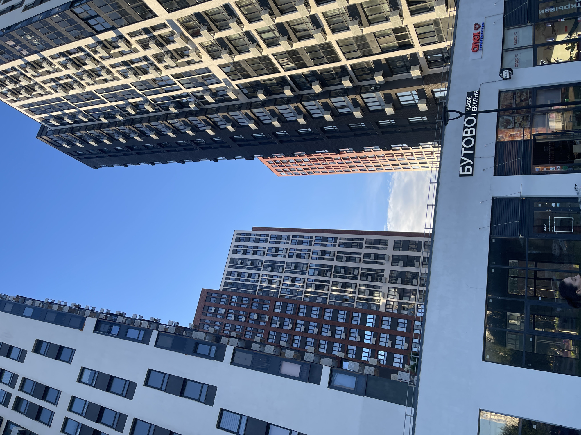 The image shows several modern residential buildings against a clear blue sky. The buildings are tall and have a grid-like pattern of windows and balconies. One building is primarily white, another is dark gray, and a third is brick-colored. In the foreground, part of a white building with dark-framed windows is visible. To the right, a white building with large glass windows and signage is present.
Visible text on the white building to the right includes "БУТОВО | КАФЕ | ЕКАРНЯ" and the logo "onex". Other smaller text is present but not clearly legible. The scene appears to be an urban residential complex. The lighting suggests it is daytime, likely late afternoon or early evening due to the angled sunlight creating shadows. The sky is mostly clear with a few wisps of clouds. The overall impression is one of new urban development. There are no people clearly visible in the image, though a silhouette of a person's head and shoulders can be seen in one of the glass windows on the right.