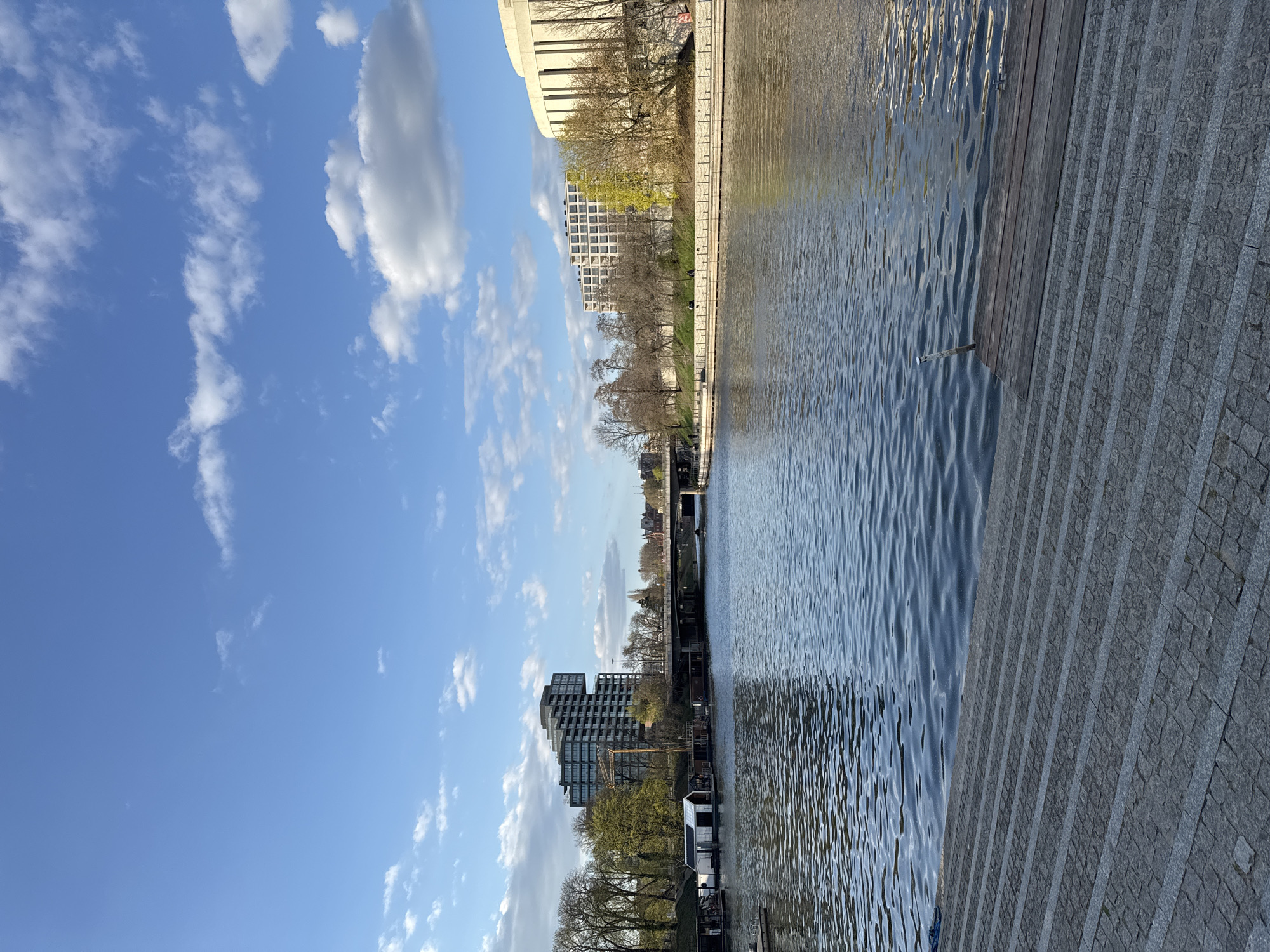 The image depicts the Brda River in Bydgoszcz, Poland, under a bright, partly cloudy sky. On the left, modern buildings line the riverbank, mixed with bare trees. Houseboats are moored along the river. A stone embankment runs along the right side. Ripples are visible on the surface of the river.