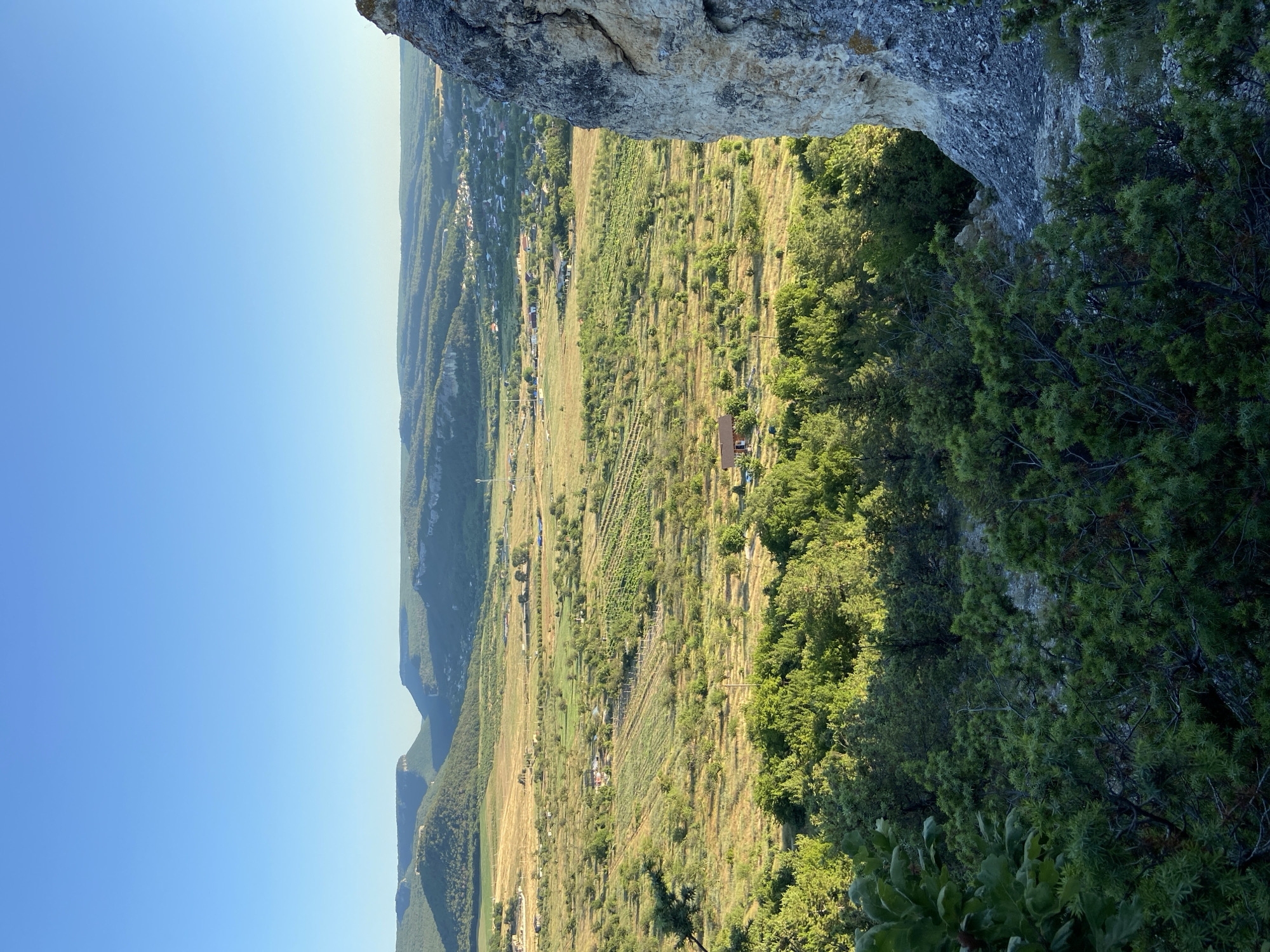 The image depicts a panoramic view of a rural landscape under a clear blue sky. In the foreground, a dense growth of green trees and shrubs occupies the bottom portion of the frame, with a large, light-colored rock formation dominating the right side. Beyond this, a valley unfolds, characterized by rolling hills, agricultural fields with visible rows of crops, and scattered trees. In the middle ground, a small village or settlement with numerous buildings is nestled amongst the greenery and fields. Further in the distance, more hills and mountains are visible, receding into the horizon under the soft light of what appears to be late afternoon or early morning. The overall impression is one of a serene, undeveloped countryside.