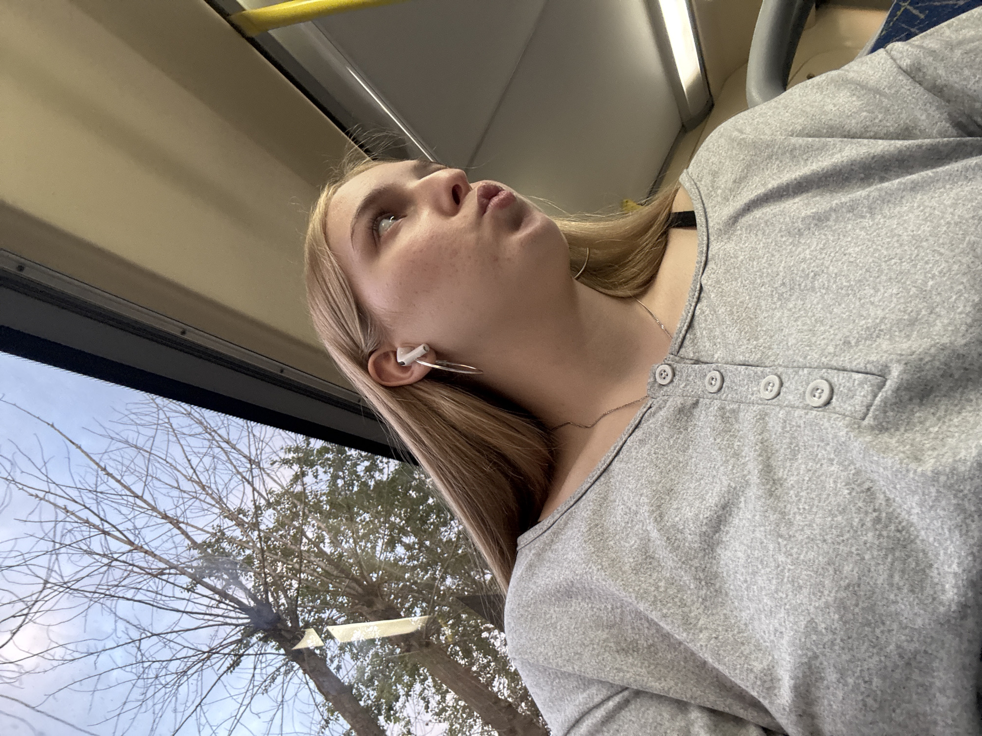 The image depicts a young blonde female, appearing to be in her late teens or early twenties, seated inside what appears to be a public transport vehicle in Perm, Russia. She is positioned in the lower-middle and right half of the frame, captured from the chest up, looking upward and to her right with her lips slightly pursed. Her long, straight blonde hair is parted on the side and falls over her shoulders. She has a fair complexion with visible pores or freckles across her face.
She is wearing a heather grey, long-sleeved Henley shirt with a V-neck and a four-button placket featuring light grey buttons. A thin silver chain necklace is visible around her neck, and a black strap, possibly a bra strap, can be seen at her left shoulder near the neckline. In her visible left ear, she wears a white wireless earbud and a large, thin silver hoop earring.
The interior of the vehicle features a light beige or off-white ceiling paneling. A bright yellow vertical grab pole is visible on the far right. A section of a blue patterned seat is partially visible in the lower right corner.
Through a large window occupying the upper and left portions of the image, bare deciduous tree branches are visible against a light blue, partly cloudy sky, suggesting a cooler season like late autumn or winter. Some green foliage is visible closer to the ground, below the bare branches. The window glass exhibits some minor streaks and reflections.