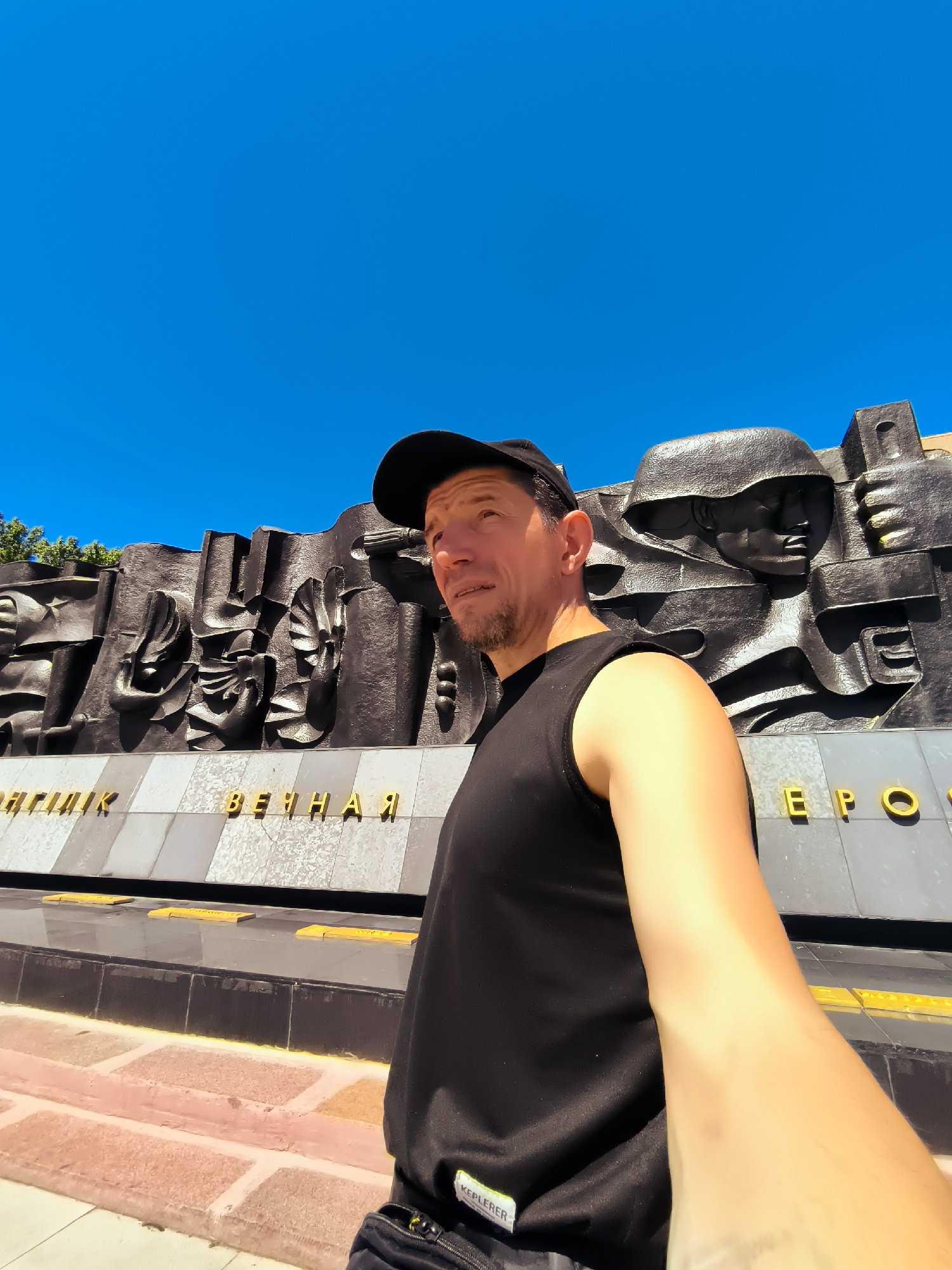 A selfie is taken from a low angle, showing a man in a black sleeveless shirt and a black baseball cap. He is looking up and to his left. Behind him is a large, dark grey monument with bas-relief sculptures of soldiers' faces and helmets. The monument has gold lettering. The sky above is clear and bright blue. The ground in front of the monument is paved with reddish-pink bricks. The man's right arm is visible, extended towards the camera, capturing the selfie. The monument appears to be a memorial, possibly related to a war or military history, located in the city of Qarağandı, Kazakhstan.